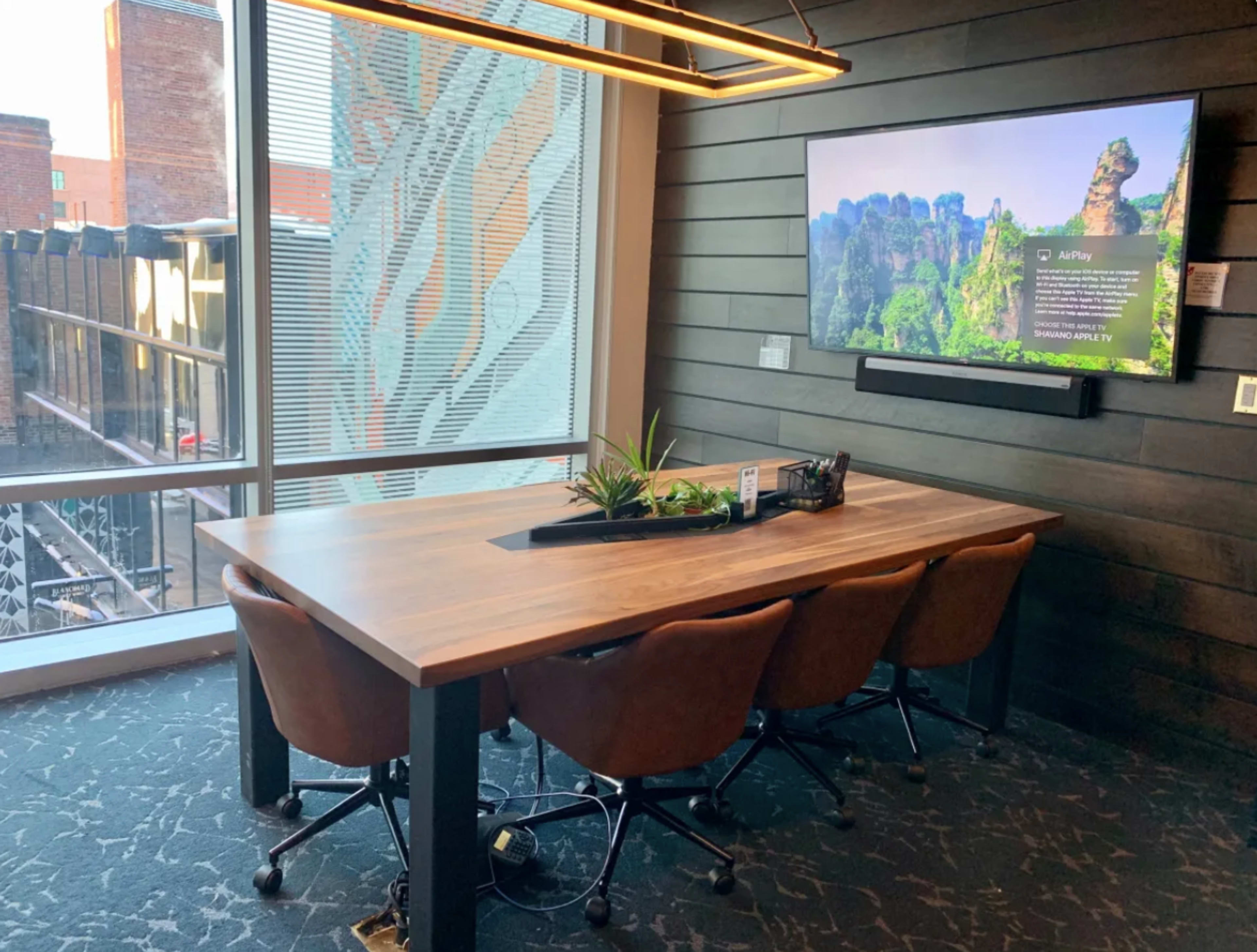 A conference table with four brown chairs is set against a window, featuring a television mounted on the wall displaying a nature scene.