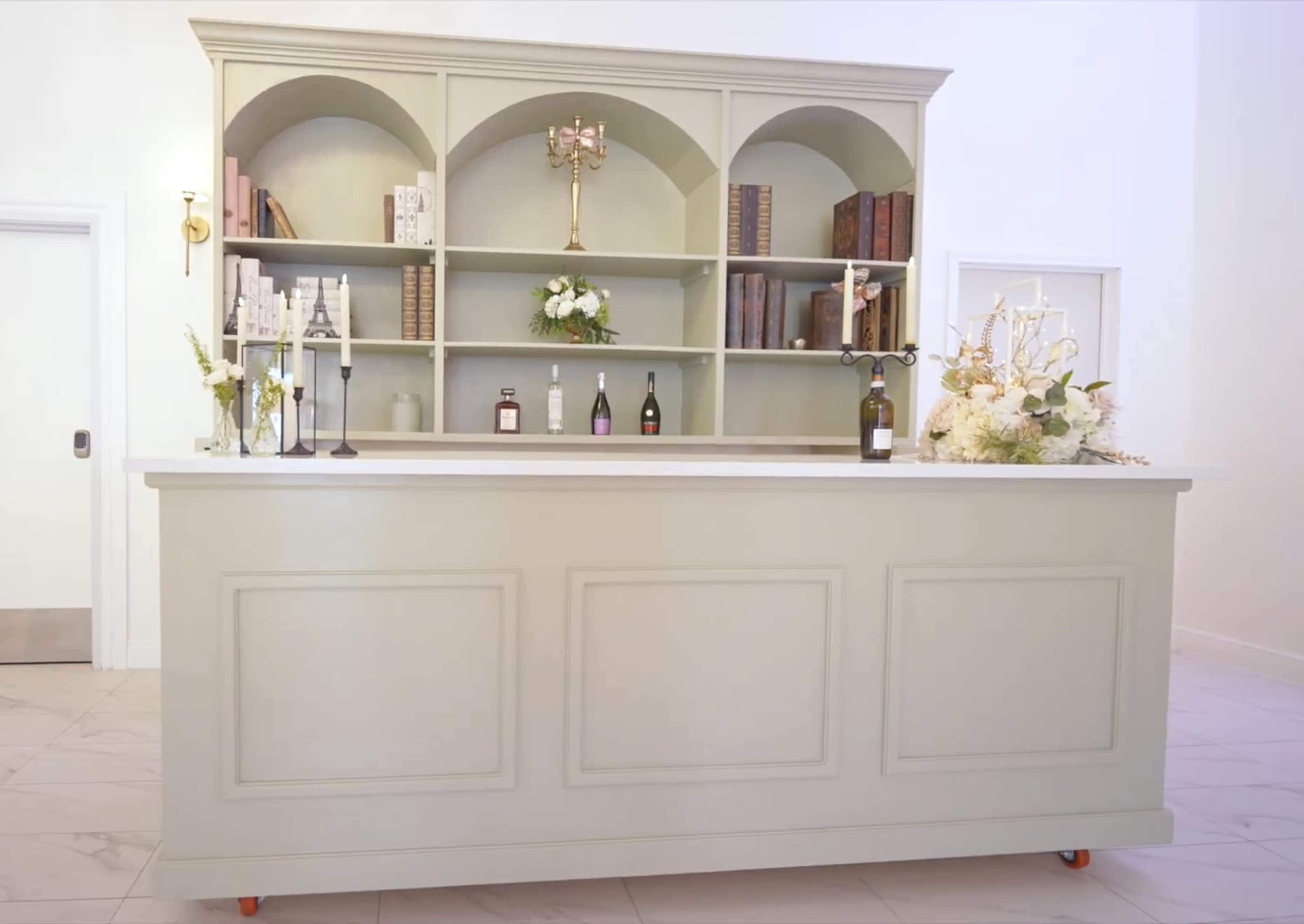 The image shows a light-colored reception desk with an arched shelving unit behind it, displaying books and floral arrangements.