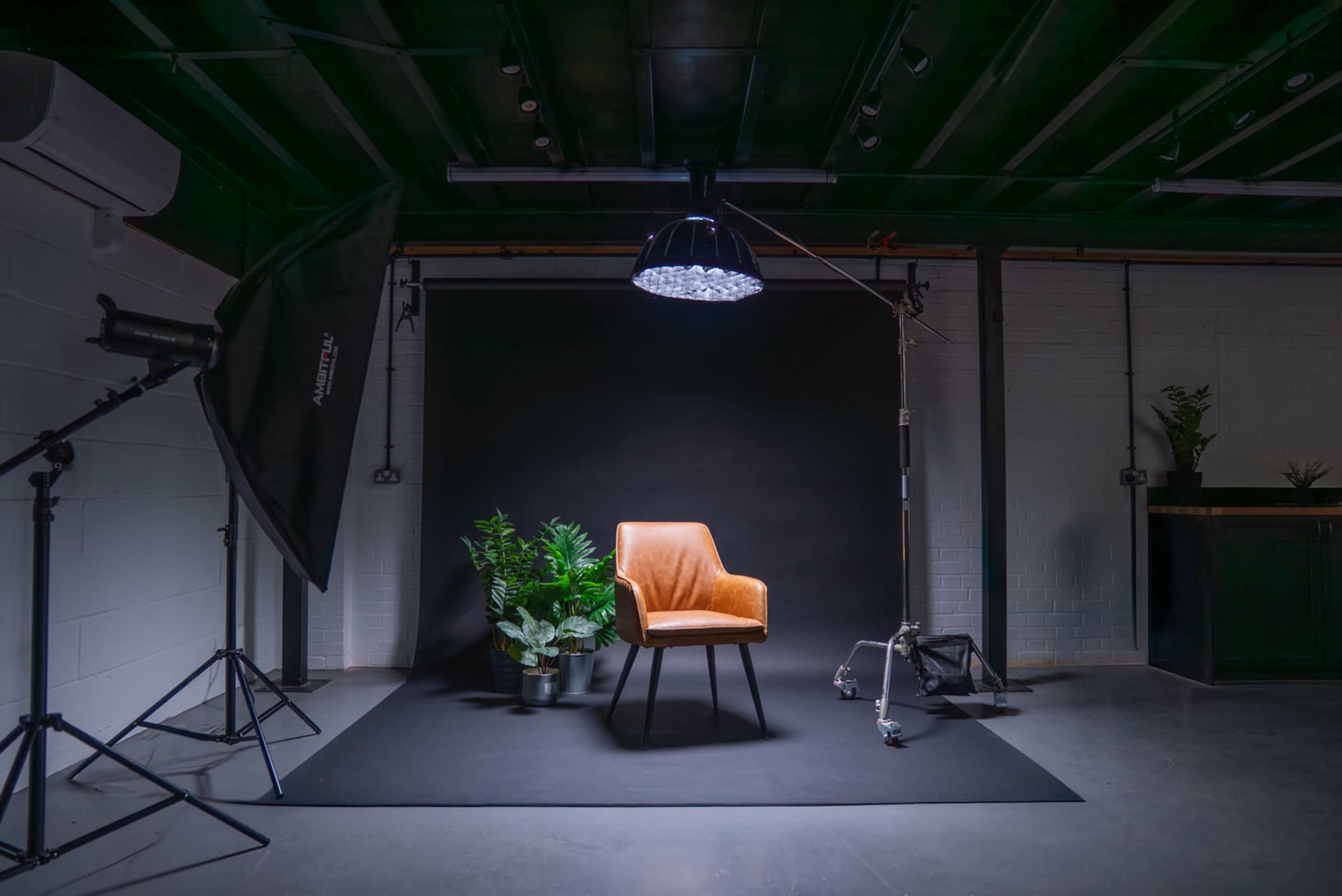 A brown chair is positioned in front of a black backdrop with potted plants nearby, illuminated by a spotlight.