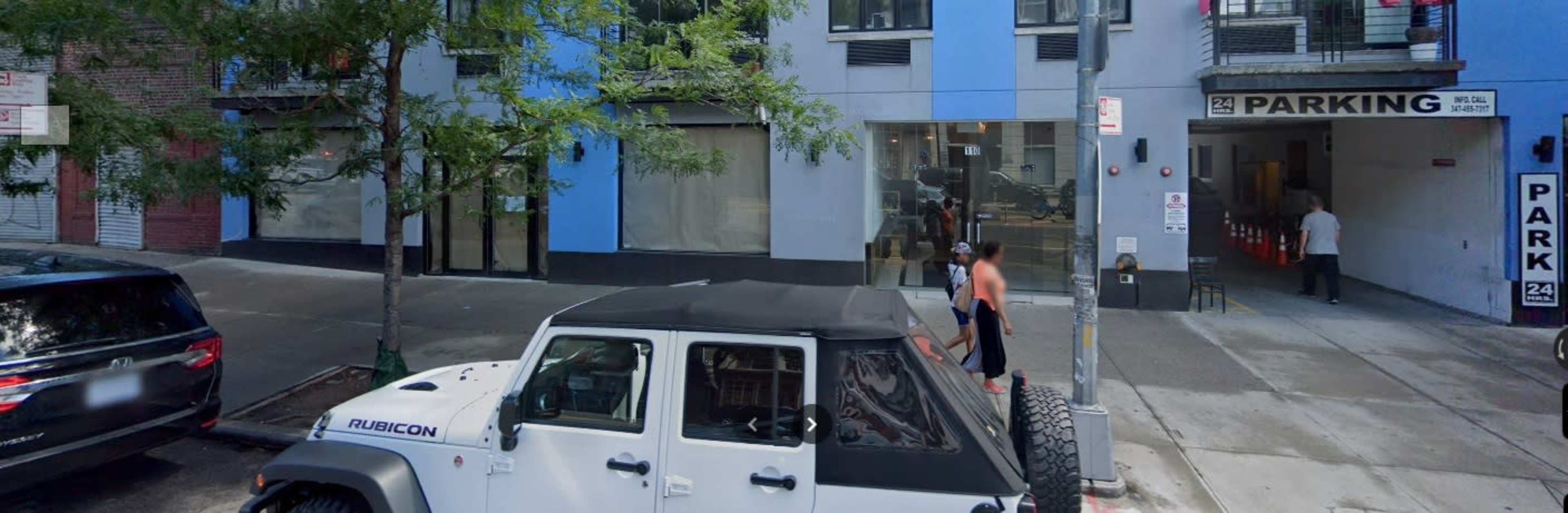 A white Jeep Rubicon is parked on the street near a blue and gray building with a sign indicating 24-hour parking.