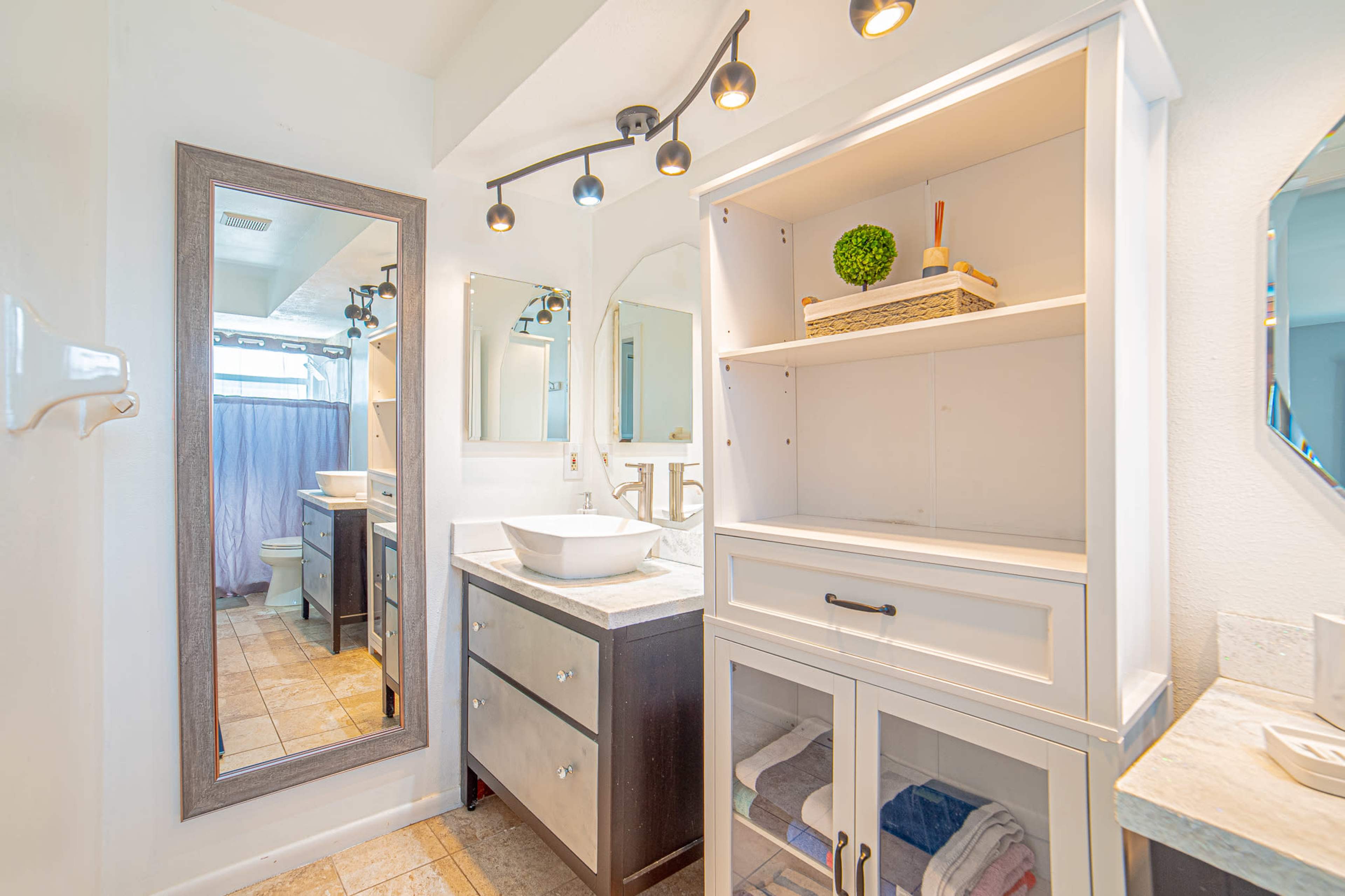 The image shows a bright bathroom featuring a white vanity with a sink, a large mirror, a wooden cabinet, and a shower area in the background.