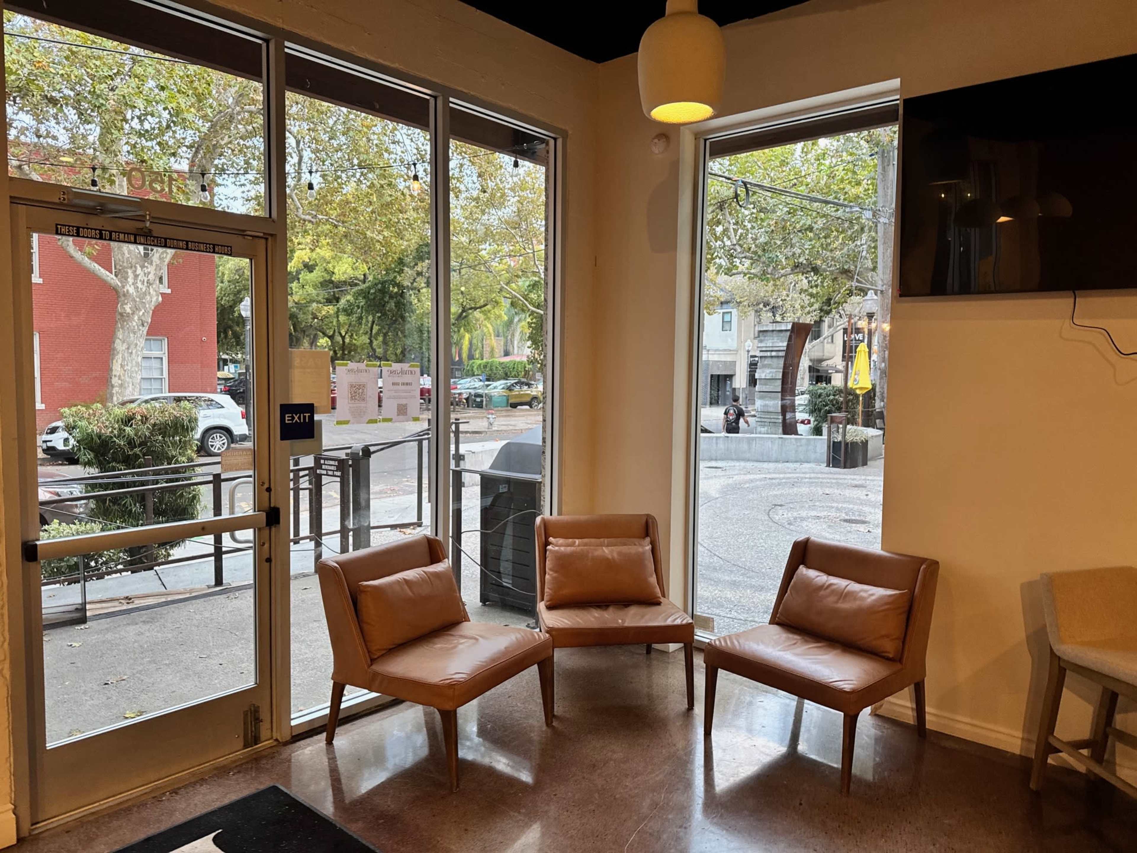The image shows a seating area with three brown chairs positioned near large windows that overlook a street outside.