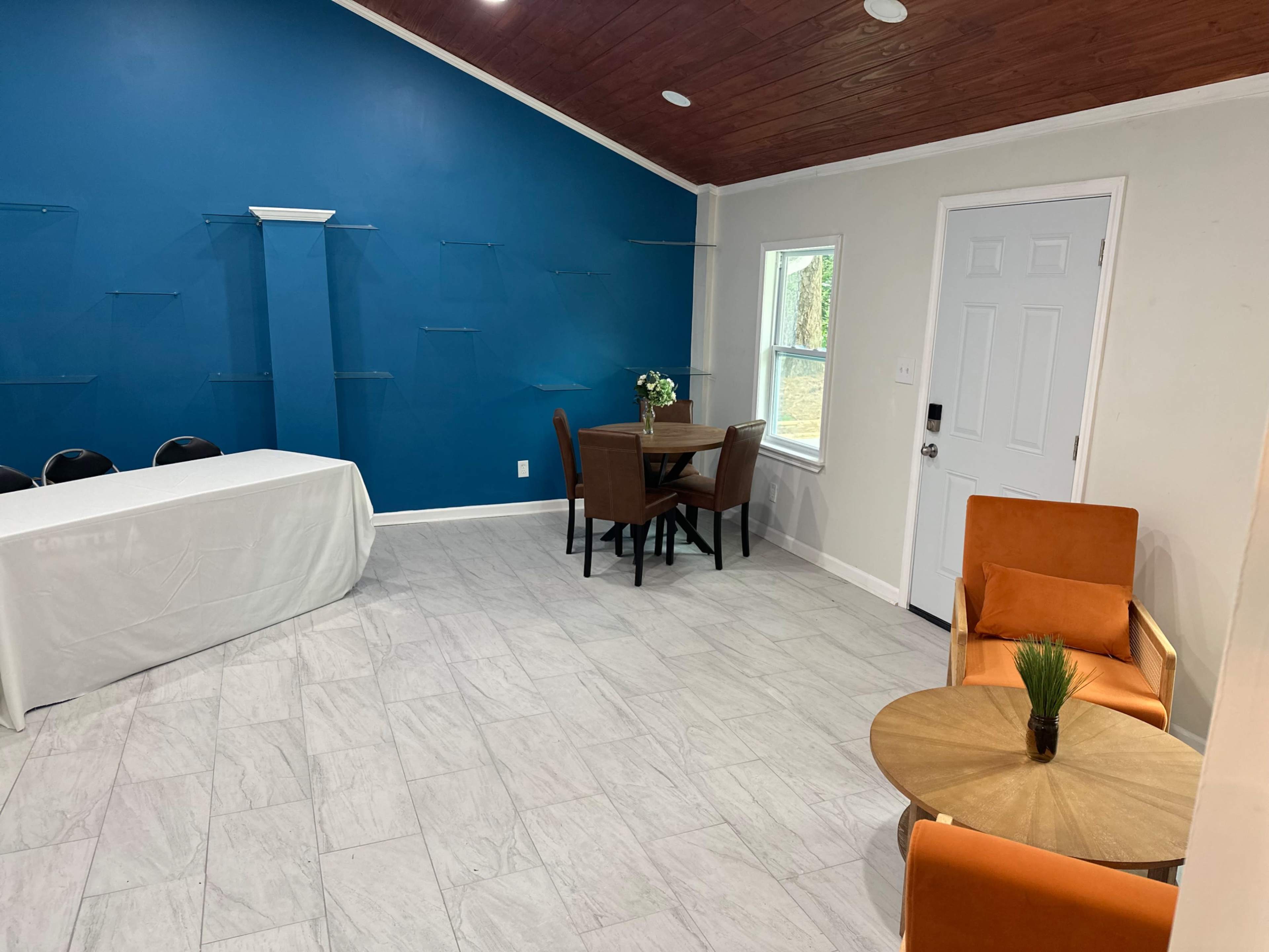 The room features a blue accent wall, a white table covered with a cloth, a dining table with chairs, and two orange armchairs next to a small round table.