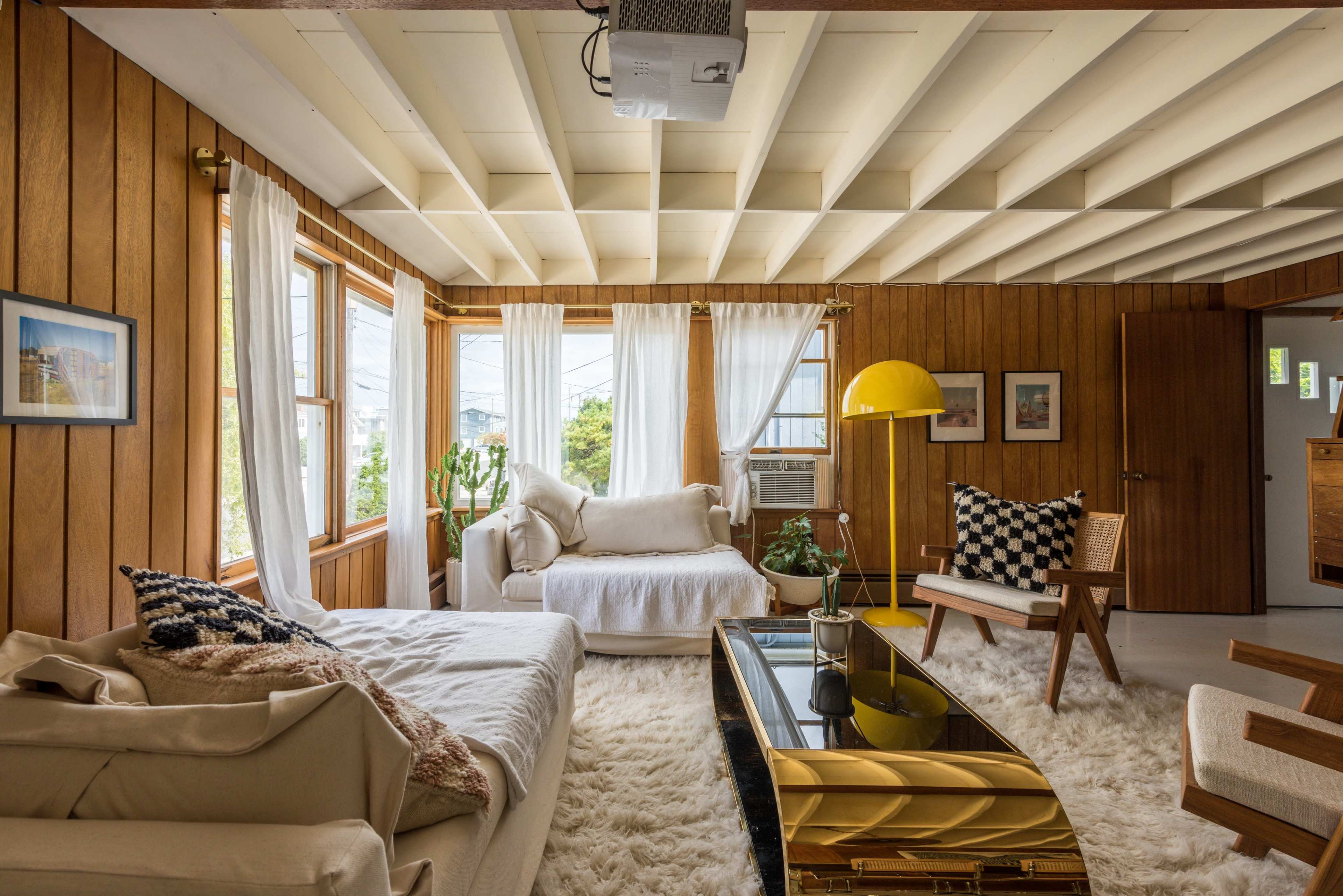 The living room features wood-paneled walls, large windows with flowing curtains, a white sofa, a yellow lamp, a glass-topped coffee table, and decorative plants.