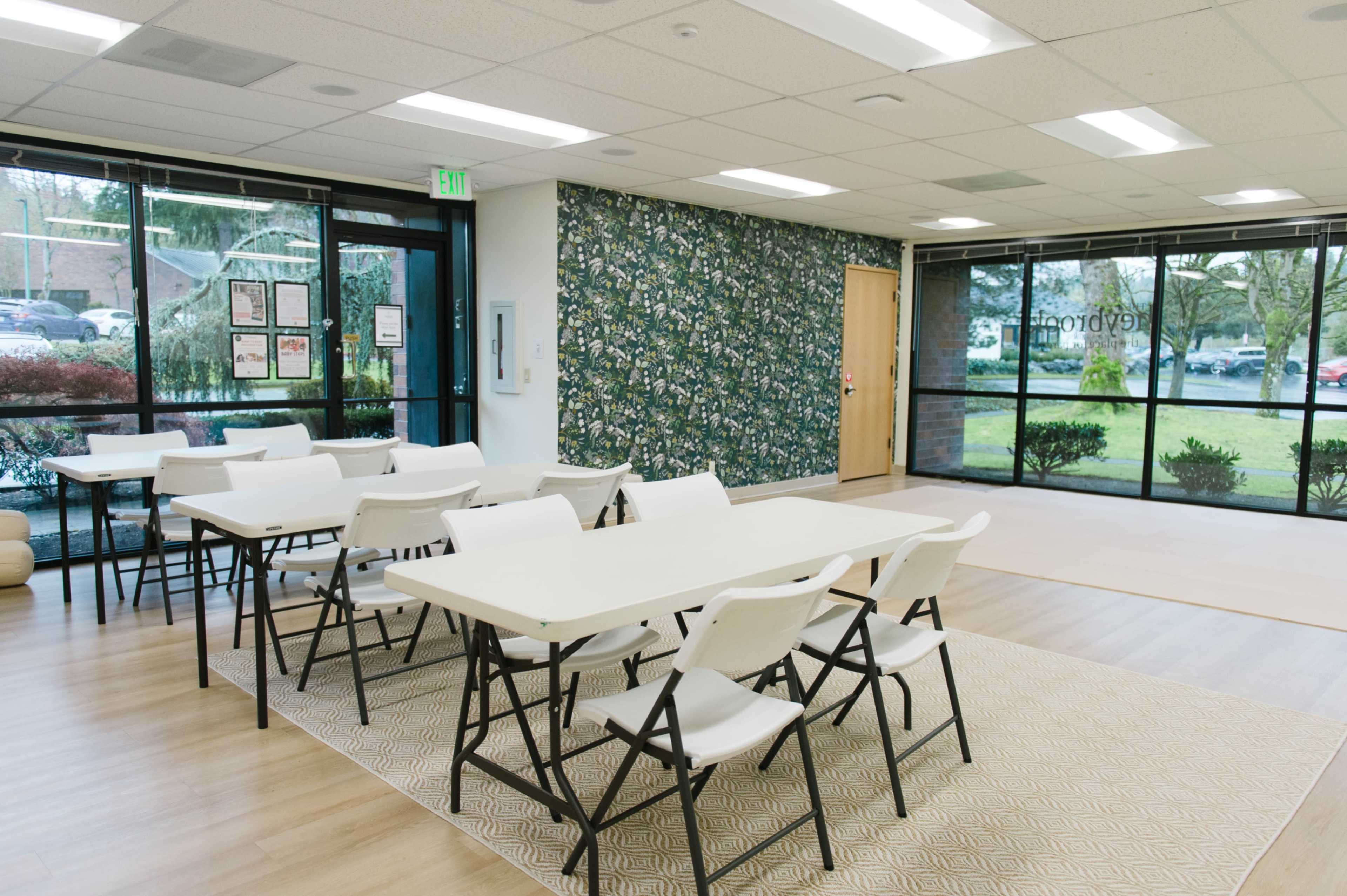 A spacious classroom features white folding tables and chairs arranged neatly on a light wooden floor, with a large window displaying greenery outside.