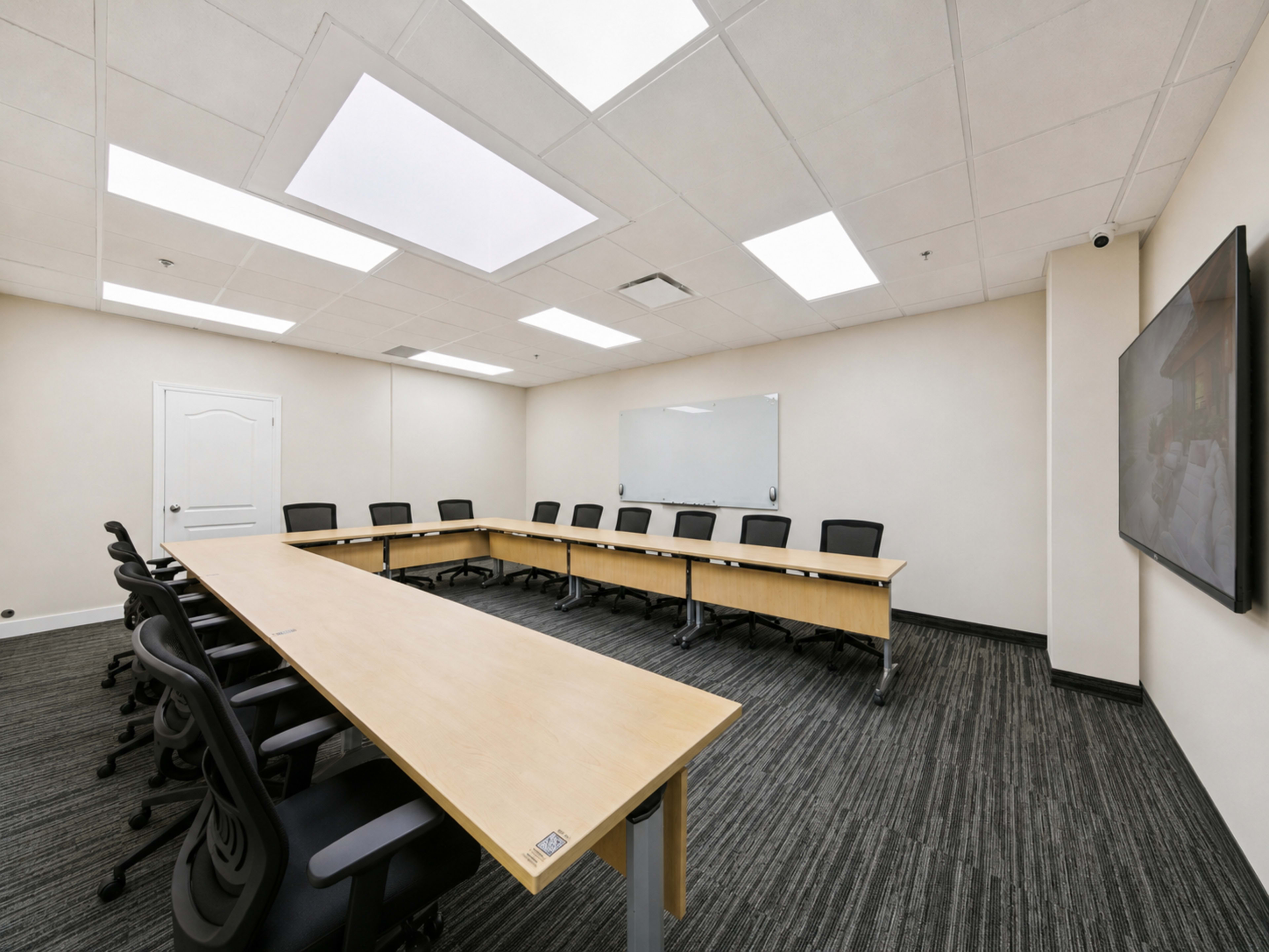 The image shows a modern conference room with a large wooden table arranged in a U-shape, black chairs, and a large wall-mounted screen.