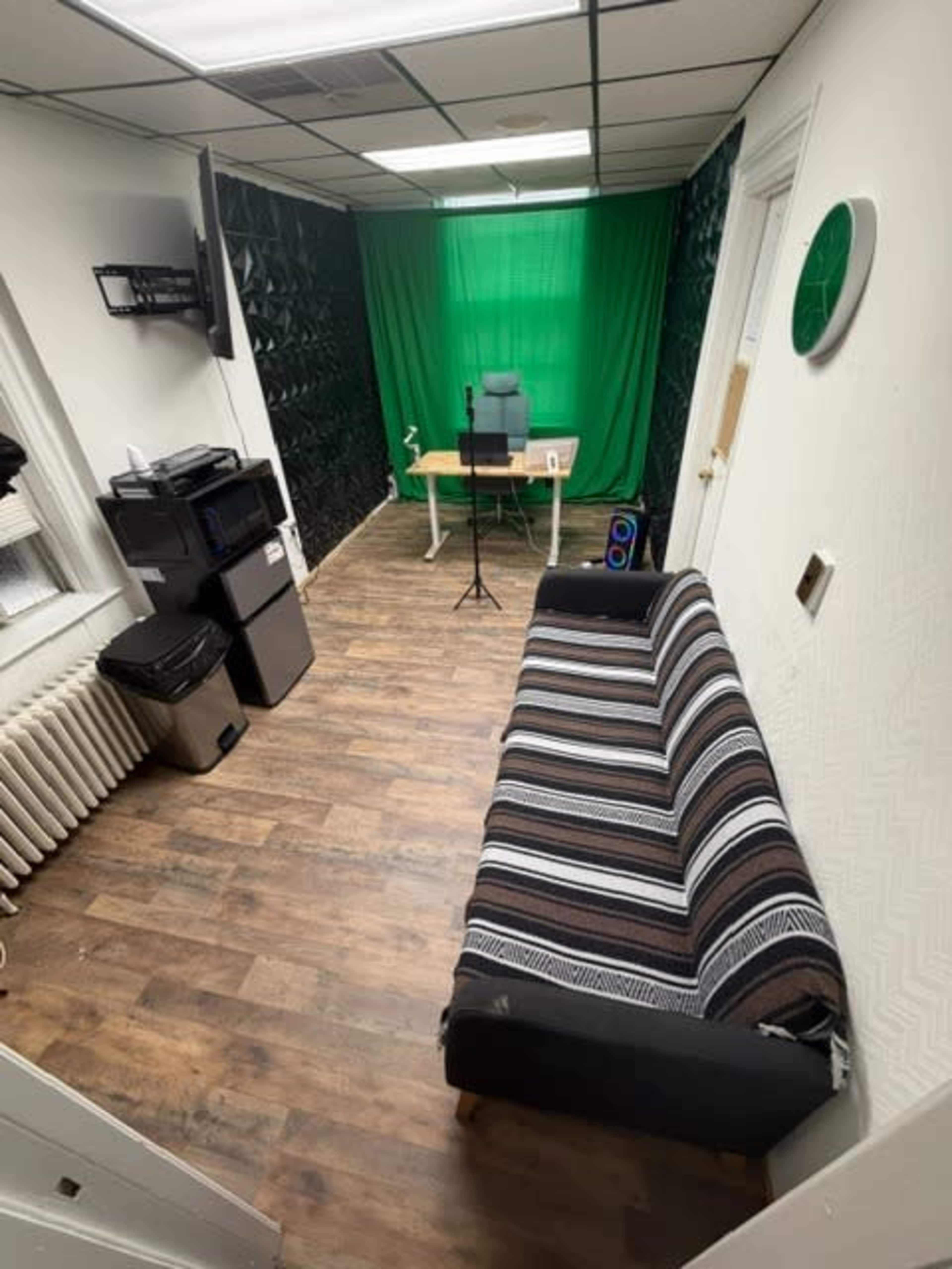 The image shows a small room with a brown wooden floor, a striped couch, a desk with a microphone and chair in front of a green backdrop, and various office equipment along one wall.
