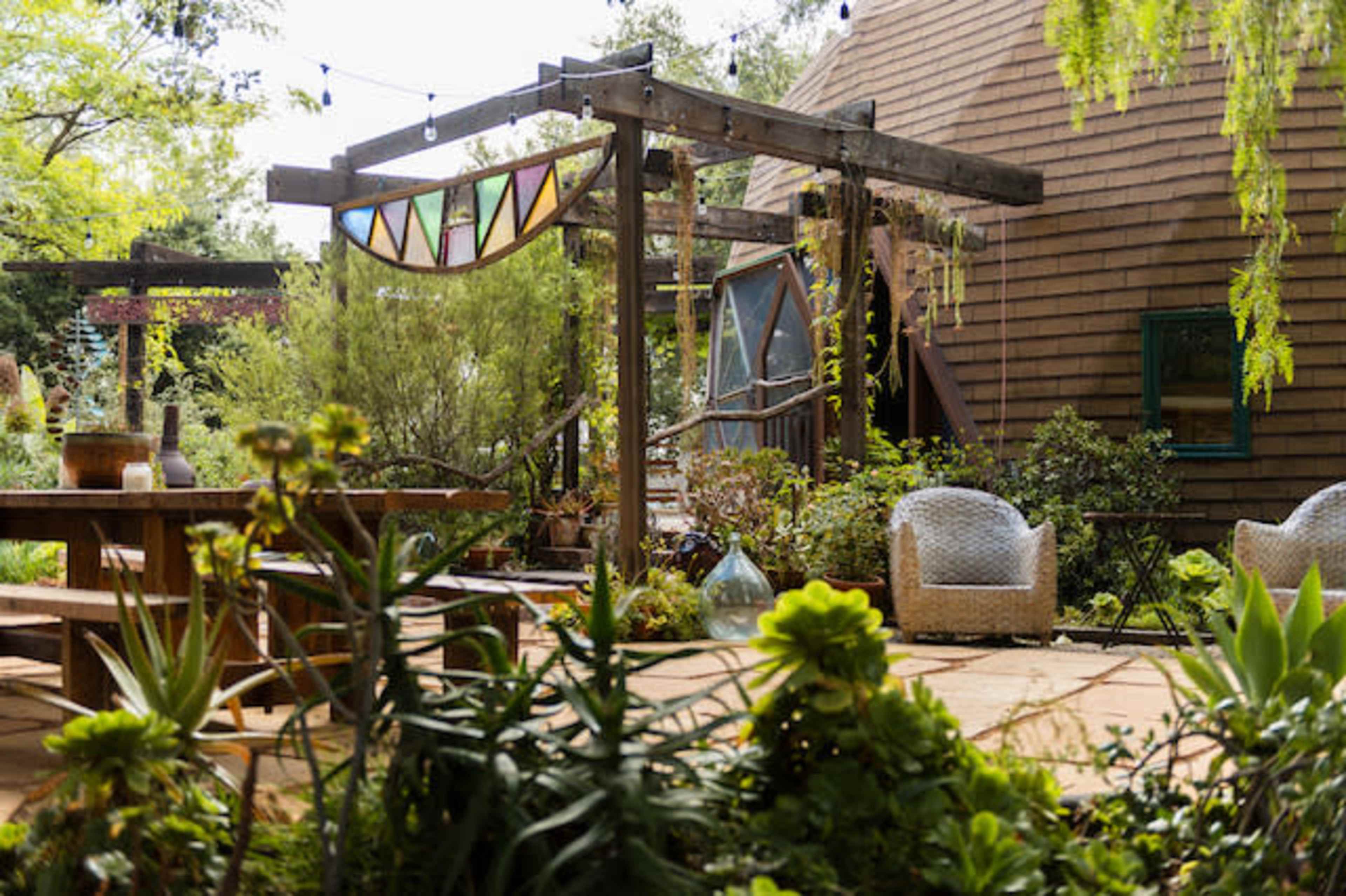 The image shows a landscaped garden area with wooden structures, a dining table, and decorative elements amid various plants and seating arrangements.