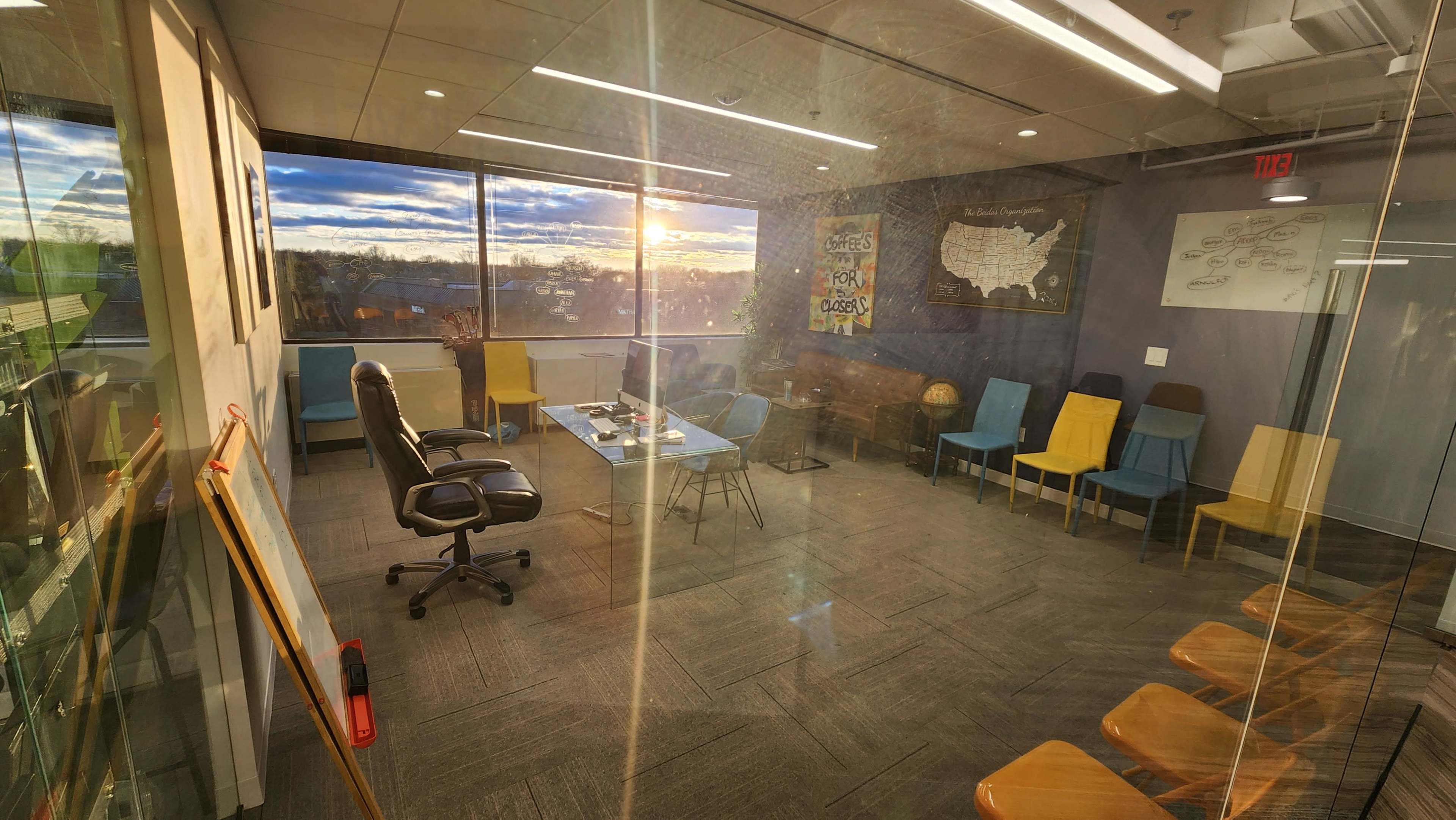 A conference room with glass walls, featuring a large window that offers a view of a sunset, and includes a black office chair, a table, and several colorful chairs arranged around the space.