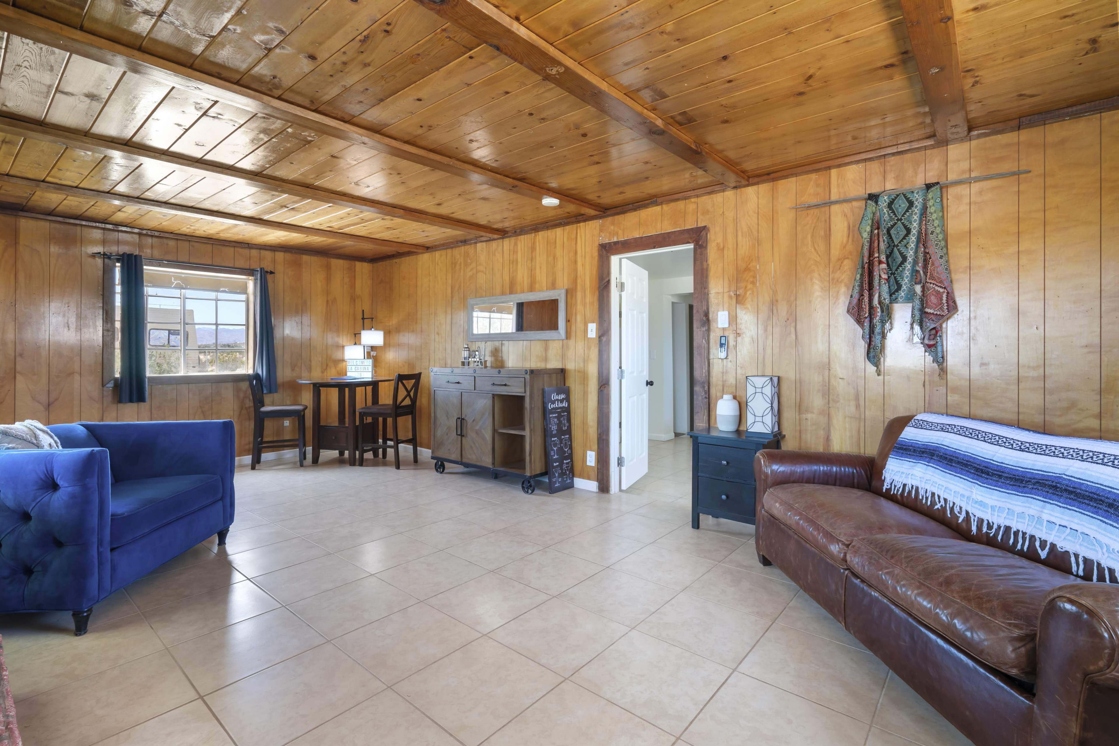 The image shows a room with wooden paneling, featuring a blue couch, a brown leather sofa, a small dining table, and a sideboard, all arranged on tiled flooring.