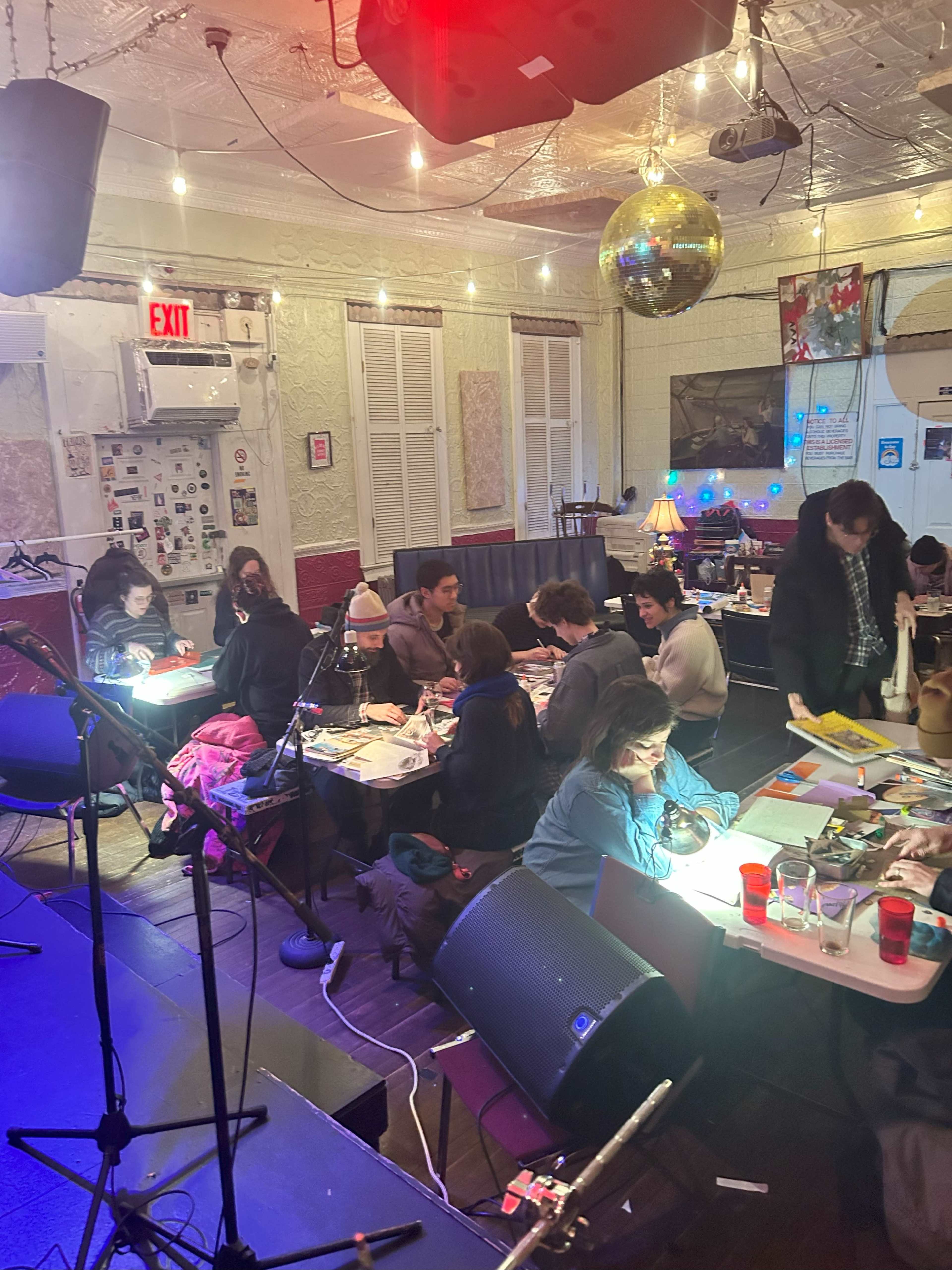 A group of people is seated at tables and on the floor of a dimly lit room, engaged in various creative activities, while a microphone and speaker sit in the foreground.