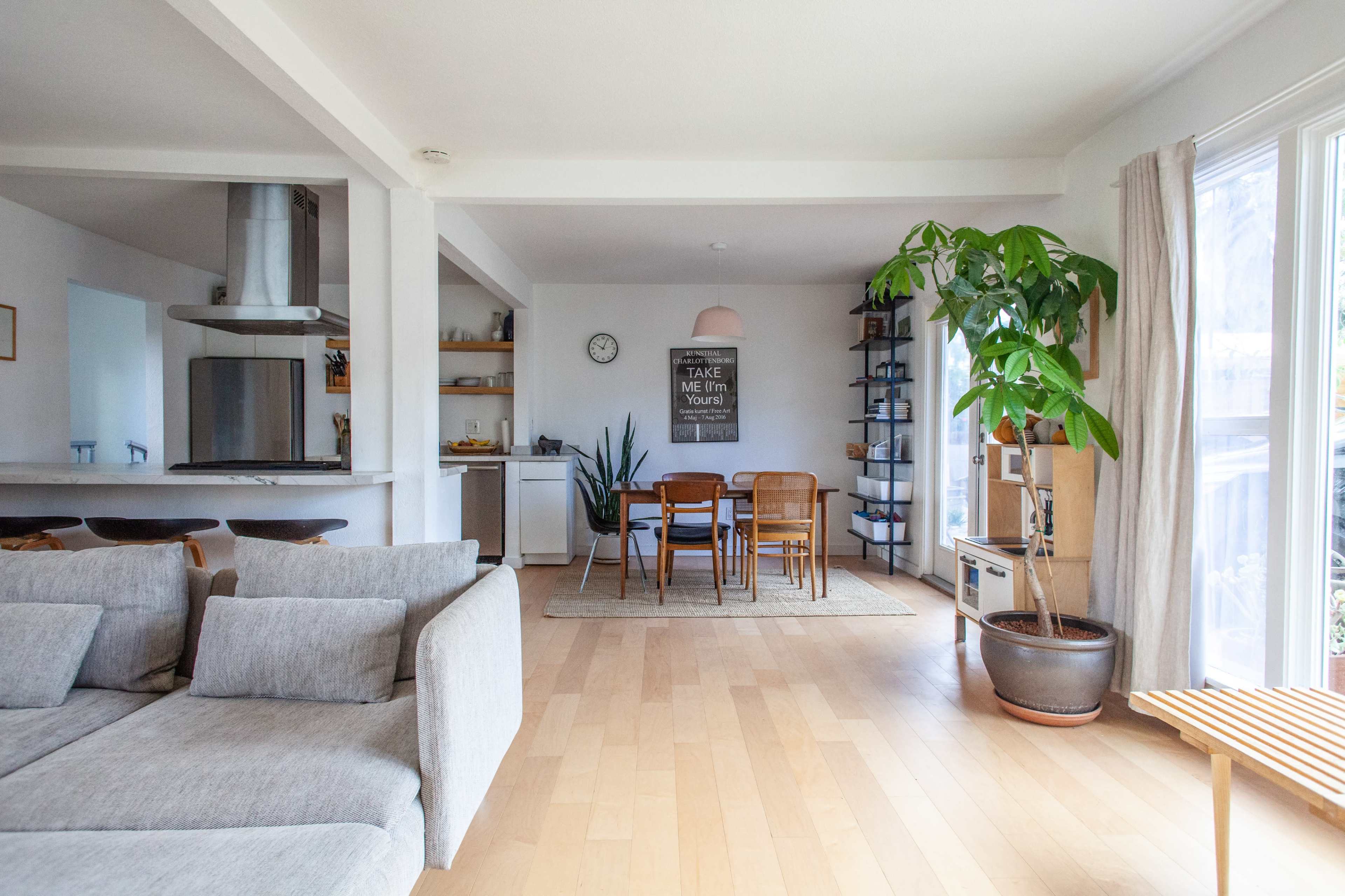A modern, open-concept living space featuring a light-colored sofa, dining area with wooden furniture, and large windows allowing natural light.