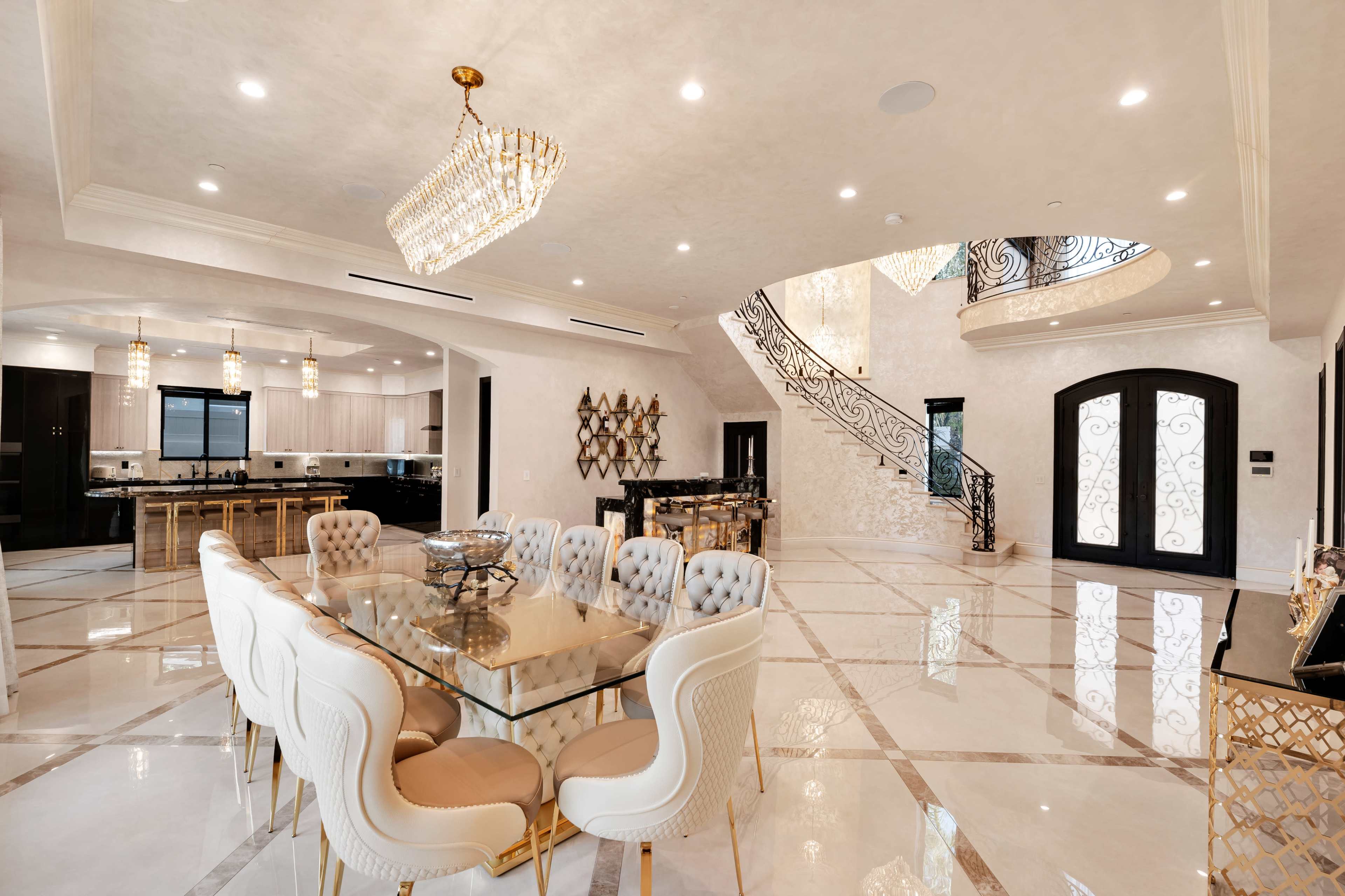 The image shows a spacious, elegantly designed dining area with a glass table surrounded by upholstered chairs, adjacent to a modern kitchen and a grand staircase.