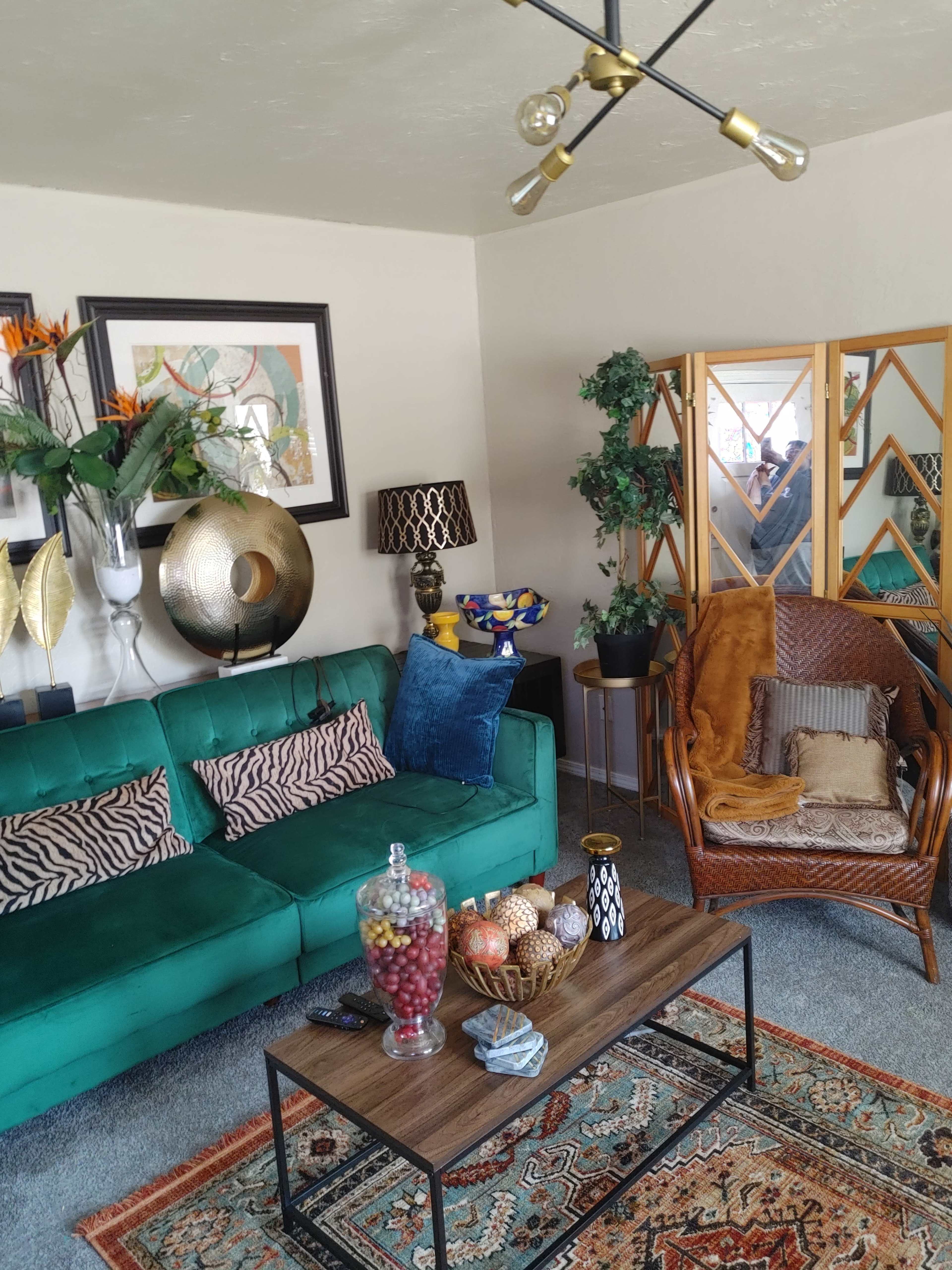 The image shows a cozy living room with a green velvet sofa, a wooden coffee table adorned with fruit, and a wicker chair beside a decorative plant and wall art.