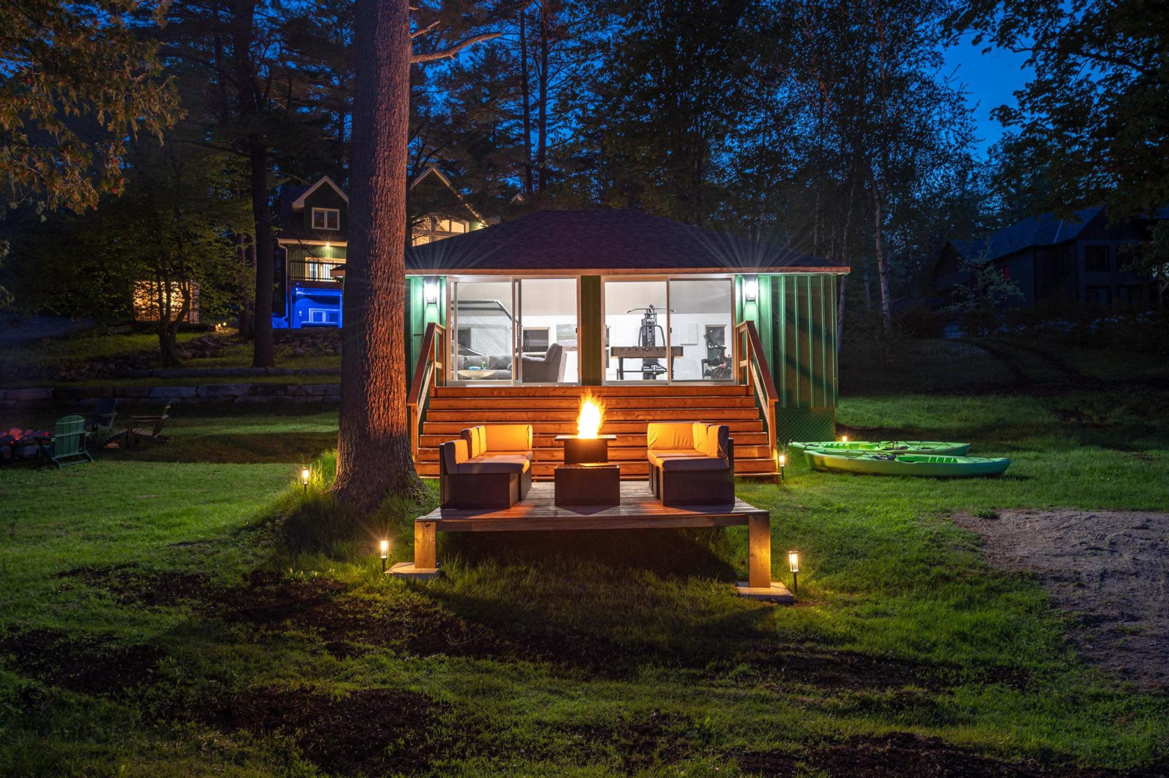A modern yurt-style cabin with steps leading to a deck and seating area is illuminated at night, surrounded by trees and a lawn, with lake houses visible in the background.