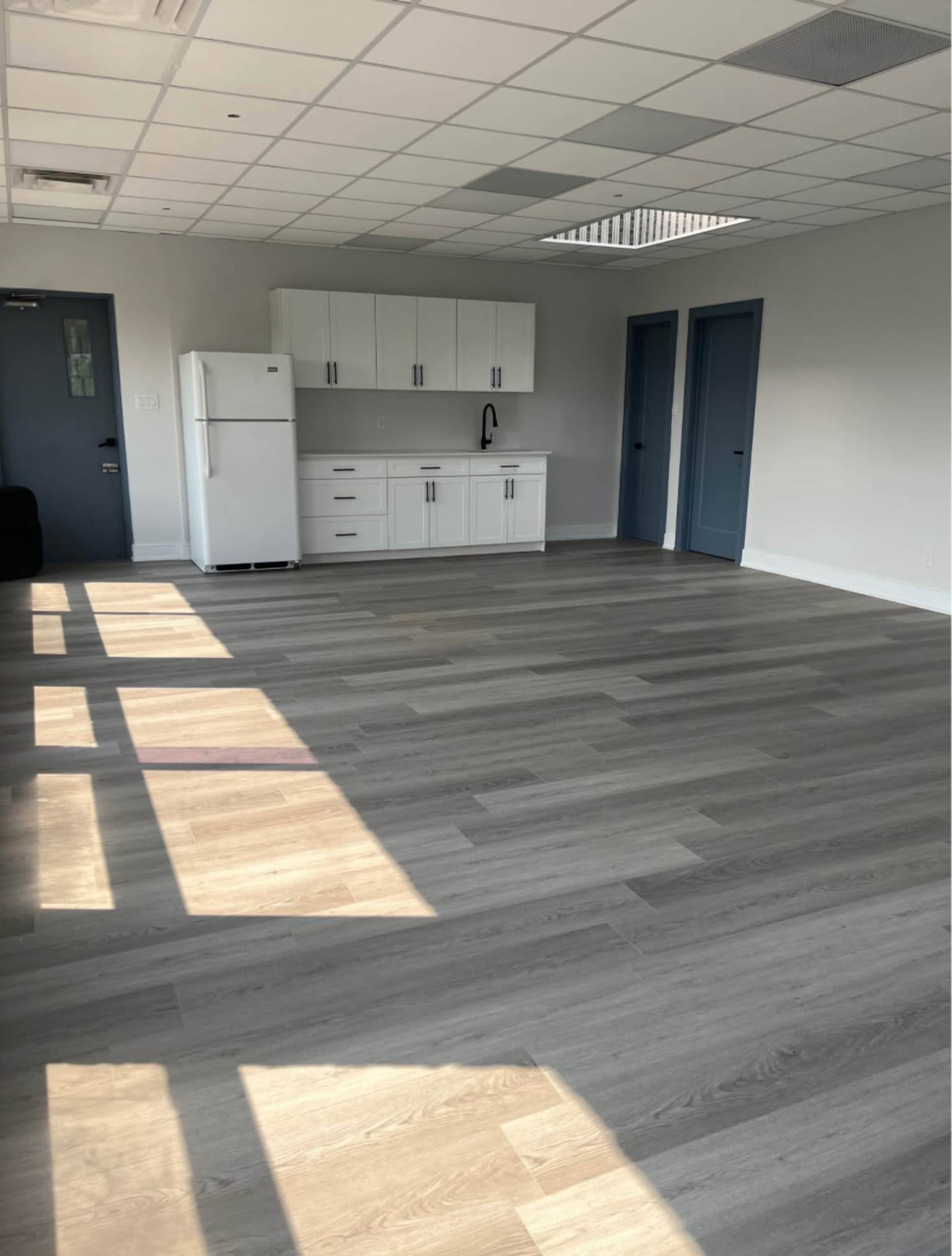 The image shows a spacious, empty room with light wood flooring, white kitchen cabinets, a refrigerator, and two blue doors.