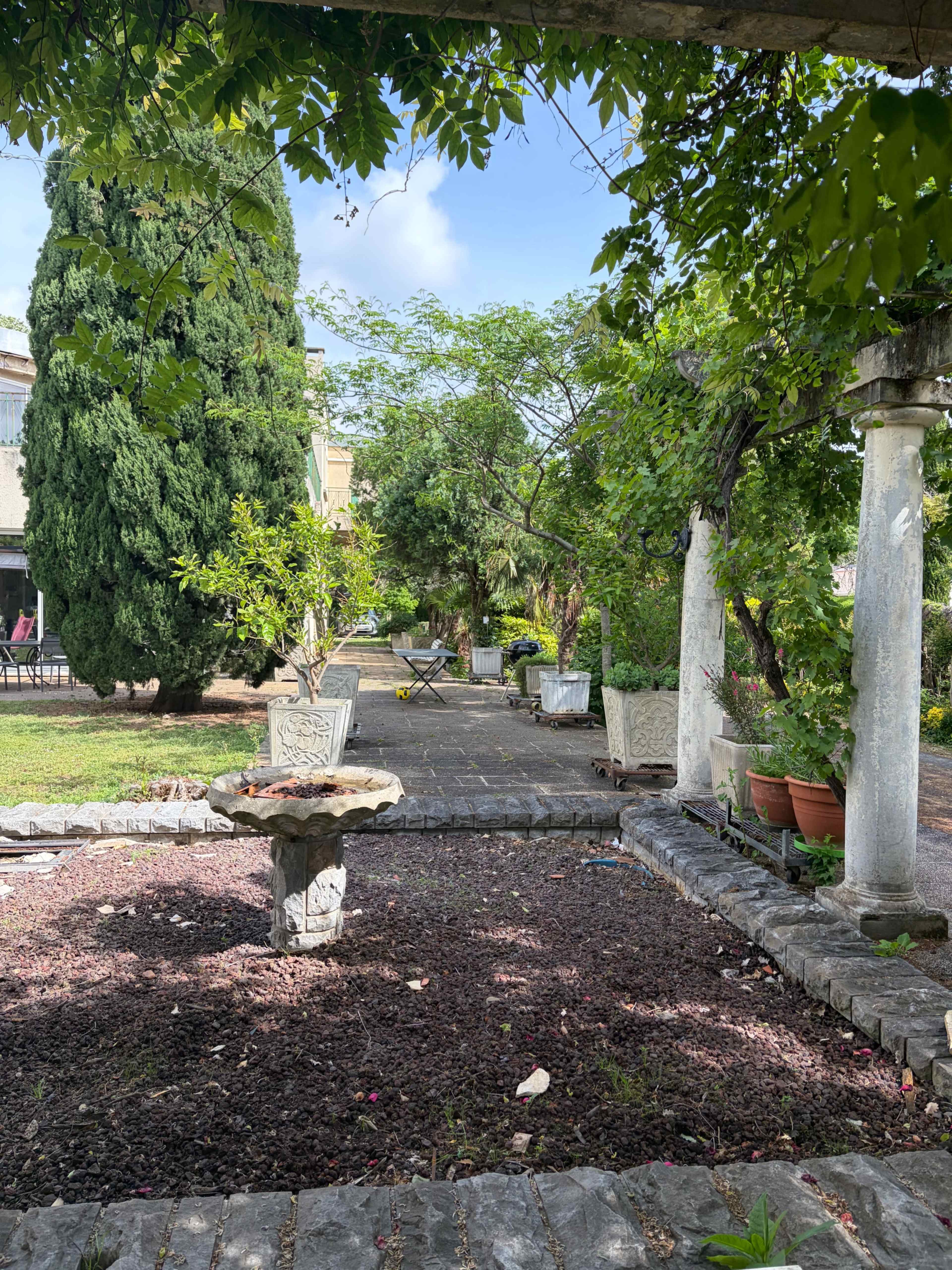 The image depicts a garden pathway lined with stone columns and surrounded by greenery, leading to a larger area in the background.