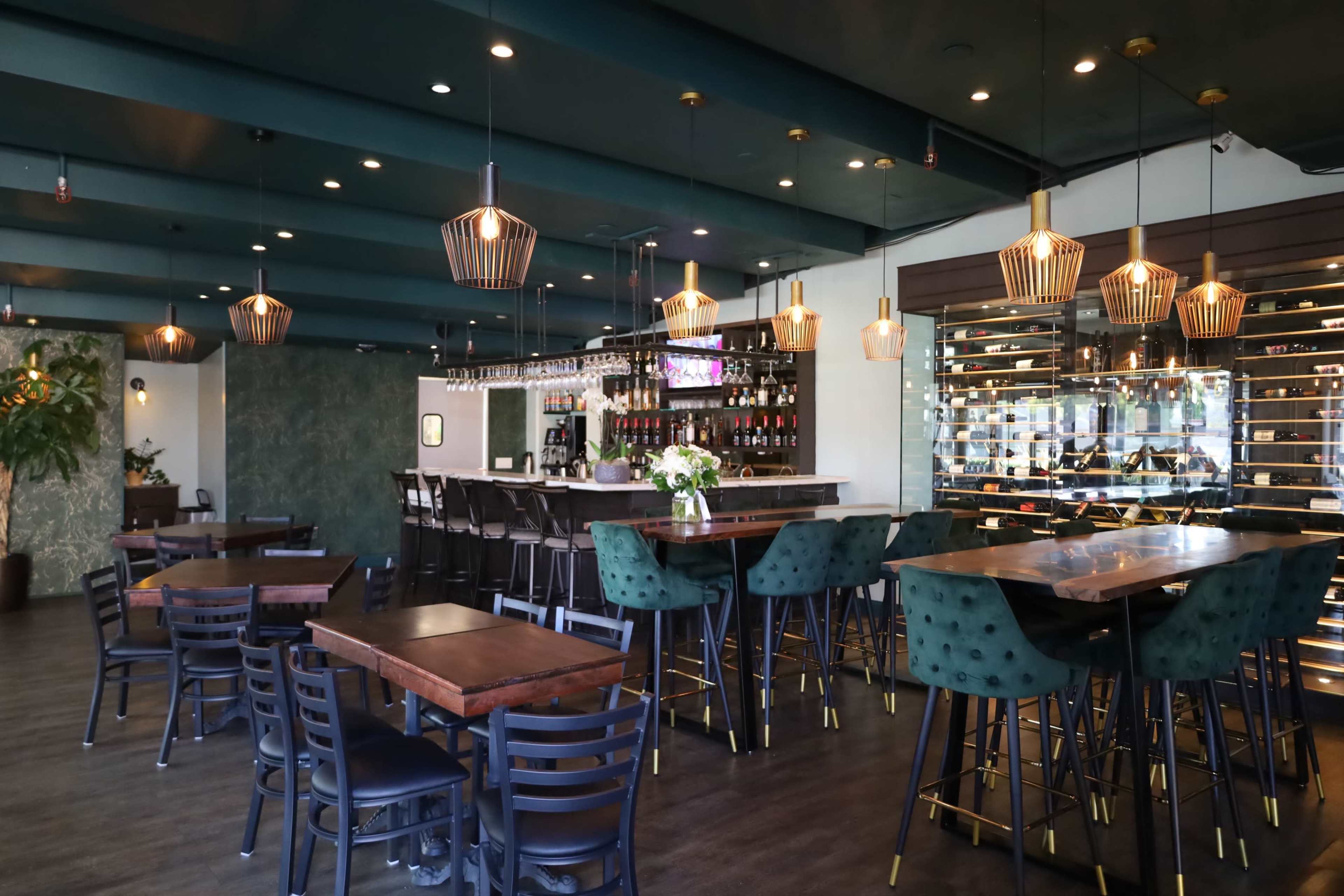 The image shows a modern bar and restaurant interior with wooden tables, green velvet chairs, and decorative pendant lighting.