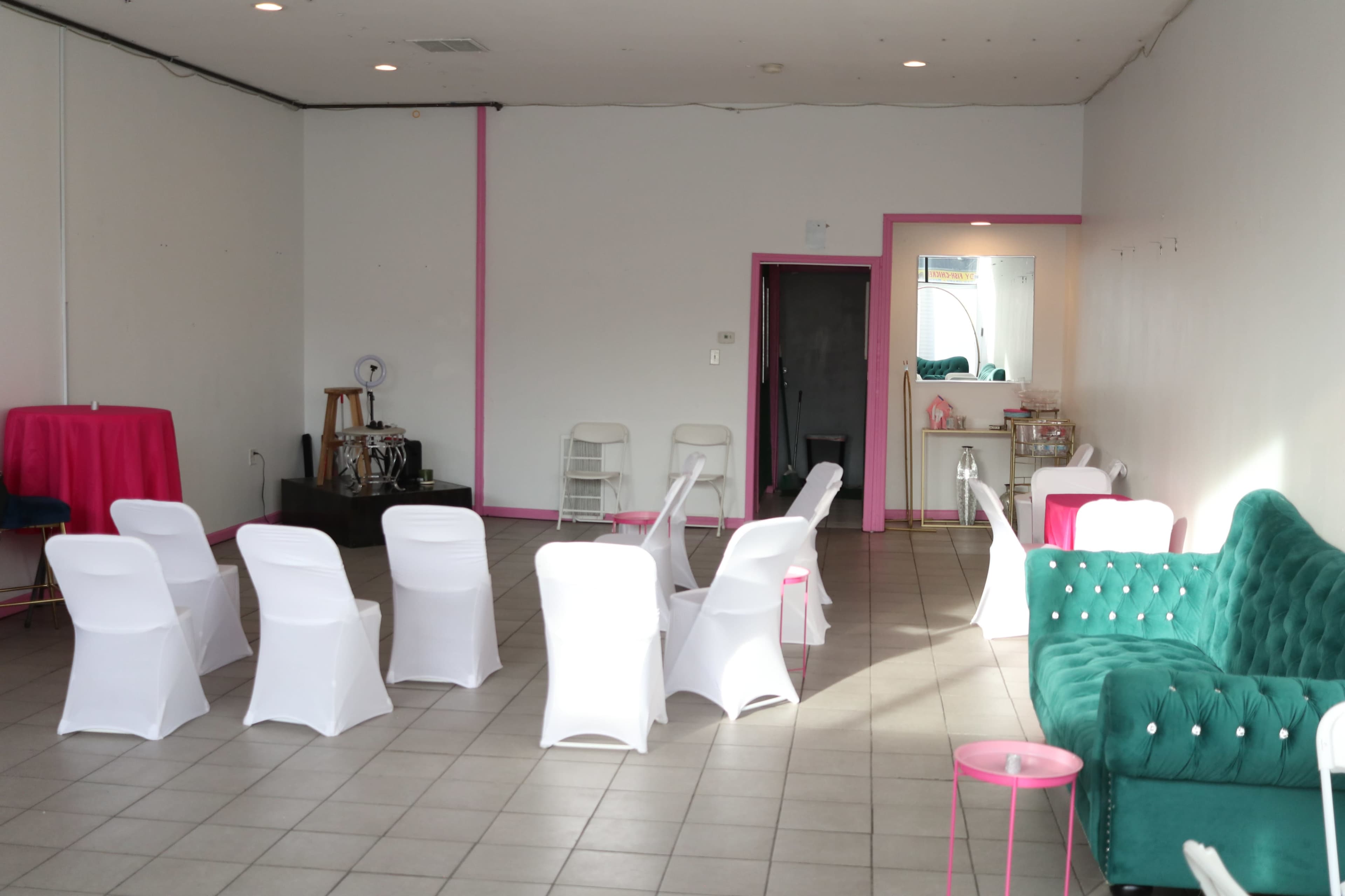 A spacious room features rows of white chairs, a green couch, and pink accents, with a small serving area in the background.
