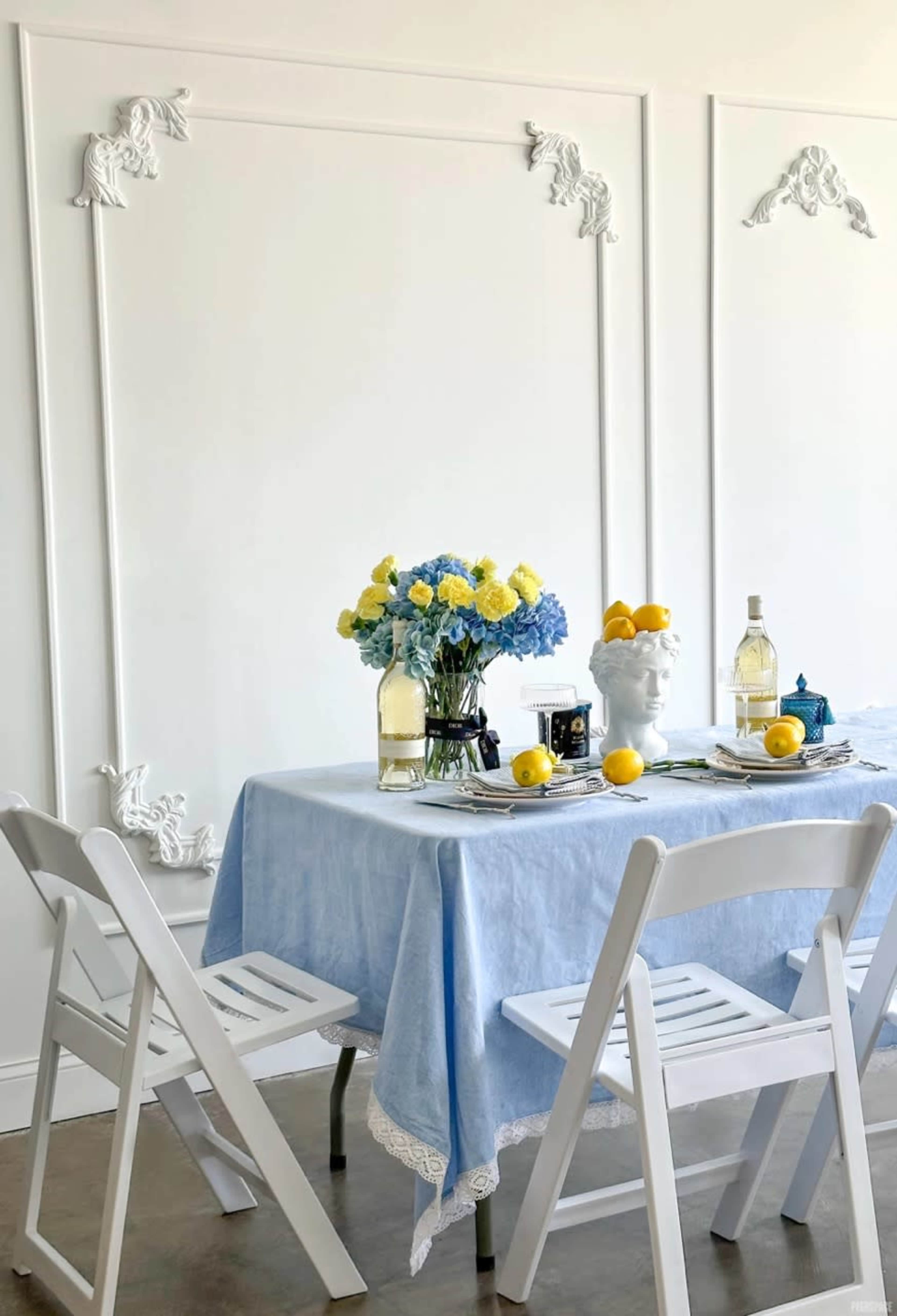A dining table is set with a blue tablecloth, a floral centerpiece, and lemons, surrounded by white chairs against a plain wall with decorative moldings.
