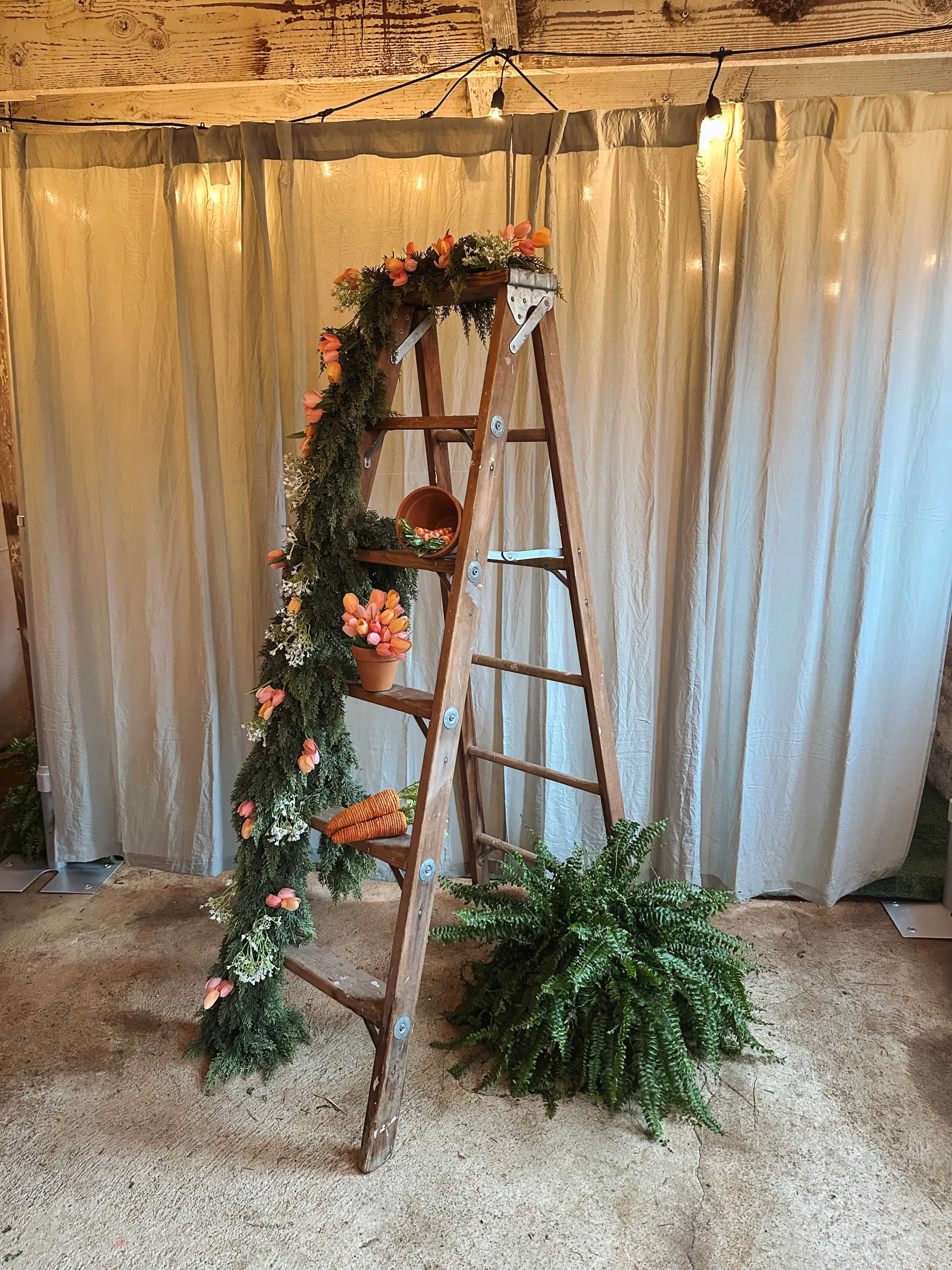 A wooden ladder decorated with a garland of flowers and potted plants stands against a backdrop of draped fabric and soft lighting.