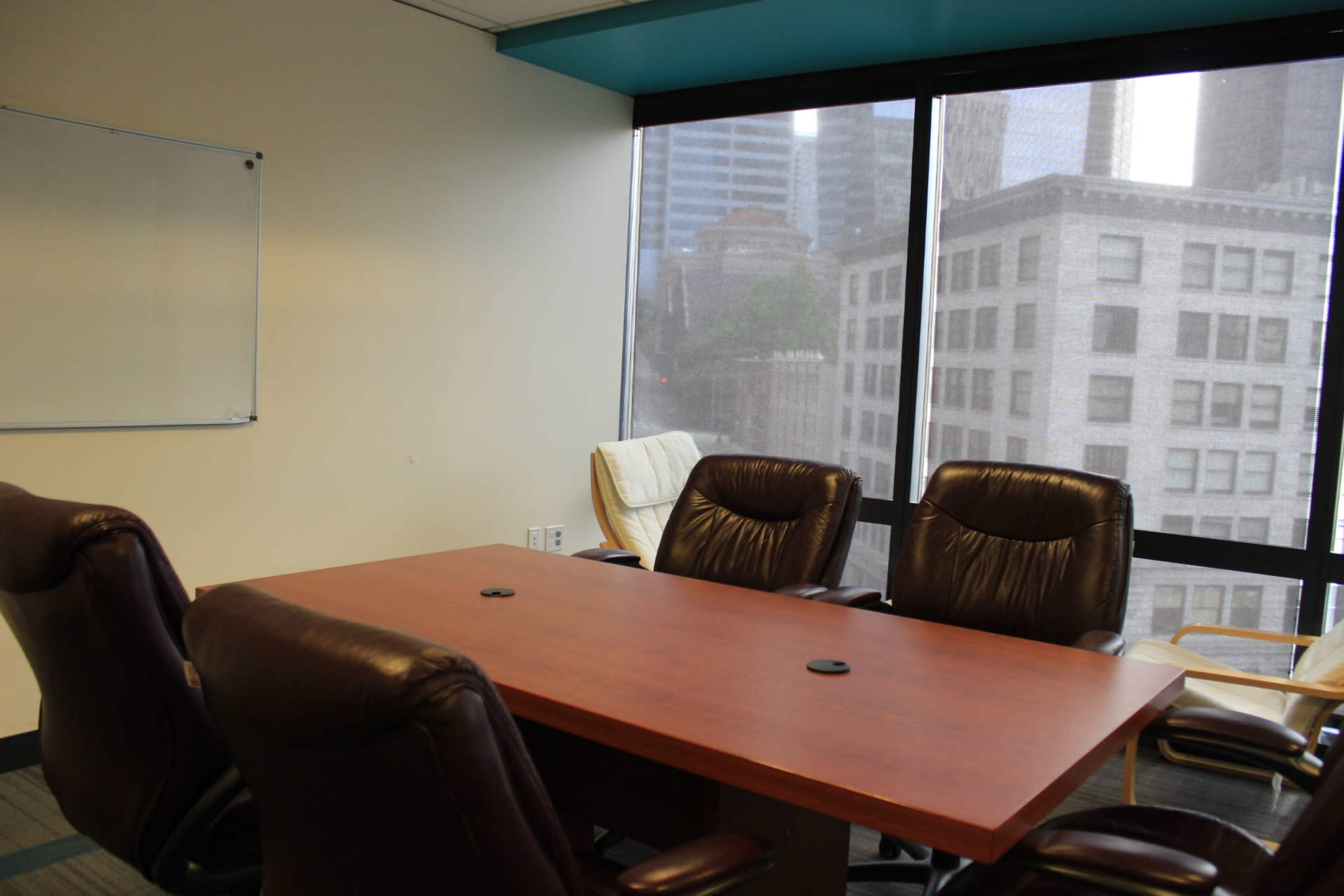 A conference room features a wooden table surrounded by leather chairs, with large windows displaying a view of tall buildings outside.