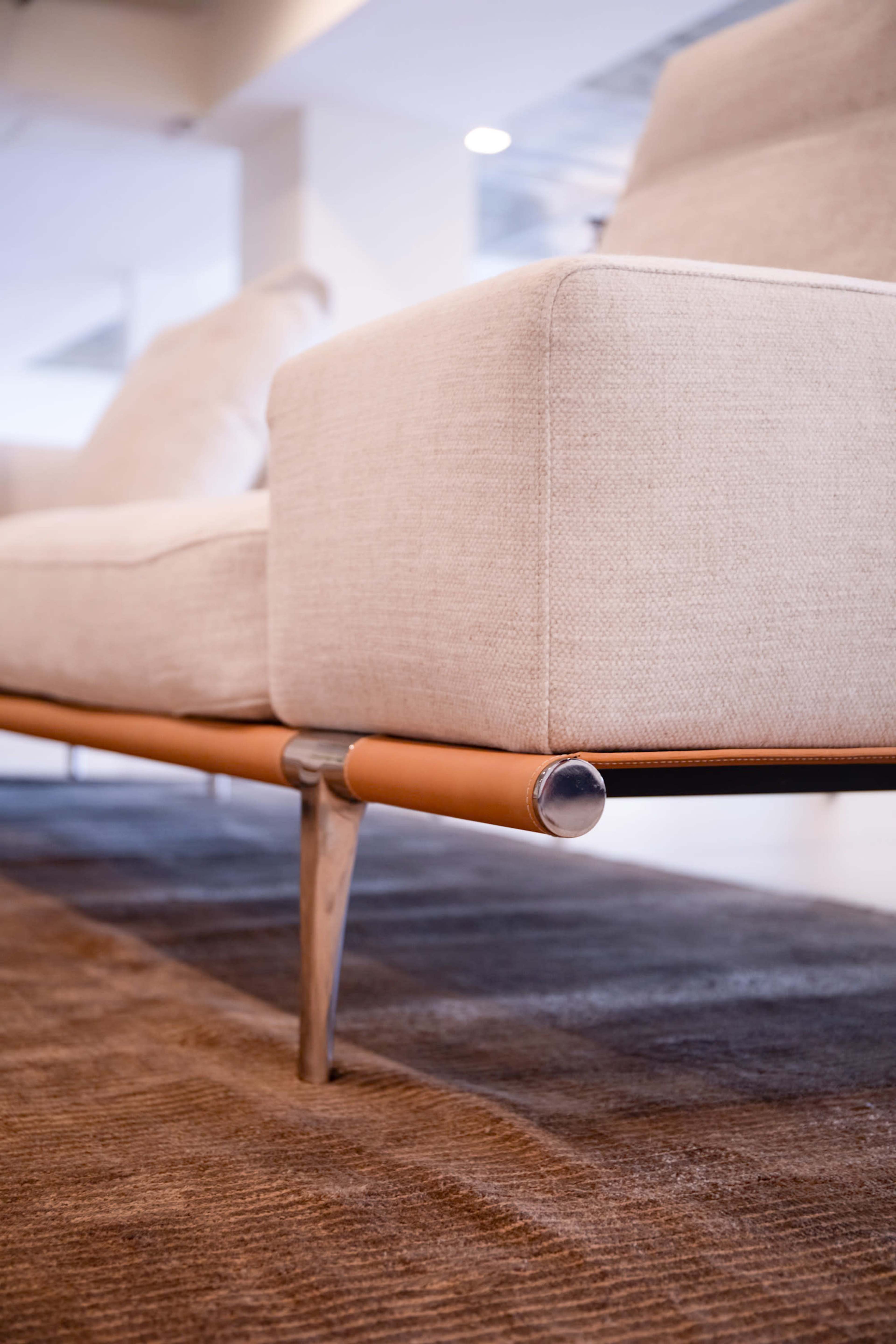 The image shows a close-up of a modern sofa with a beige fabric covering and sleek metal legs, placed on a brown rug.
