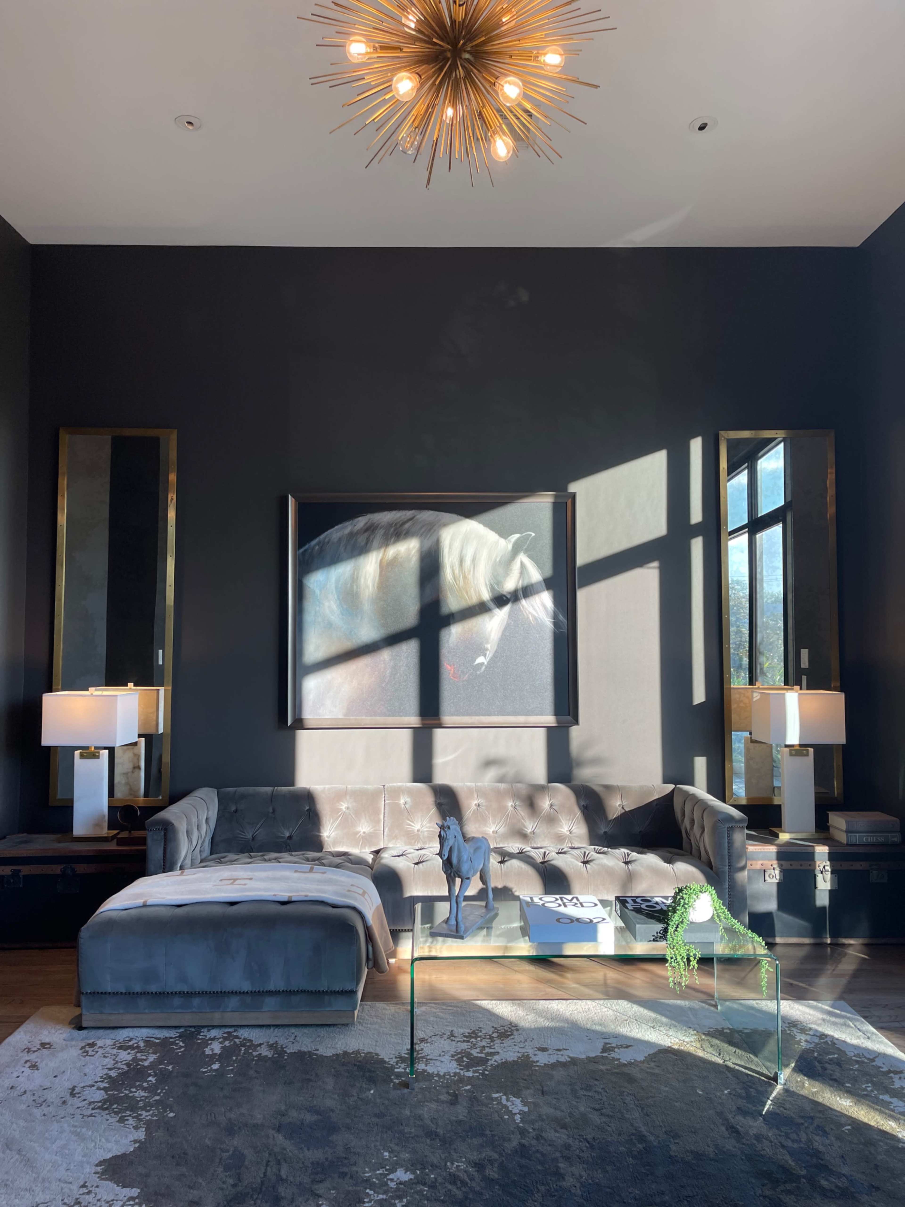 The living room features a sectional sofa, a large abstract painting, floor-to-ceiling windows, and a sunburst chandelier, with natural light casting shadows on the furniture.