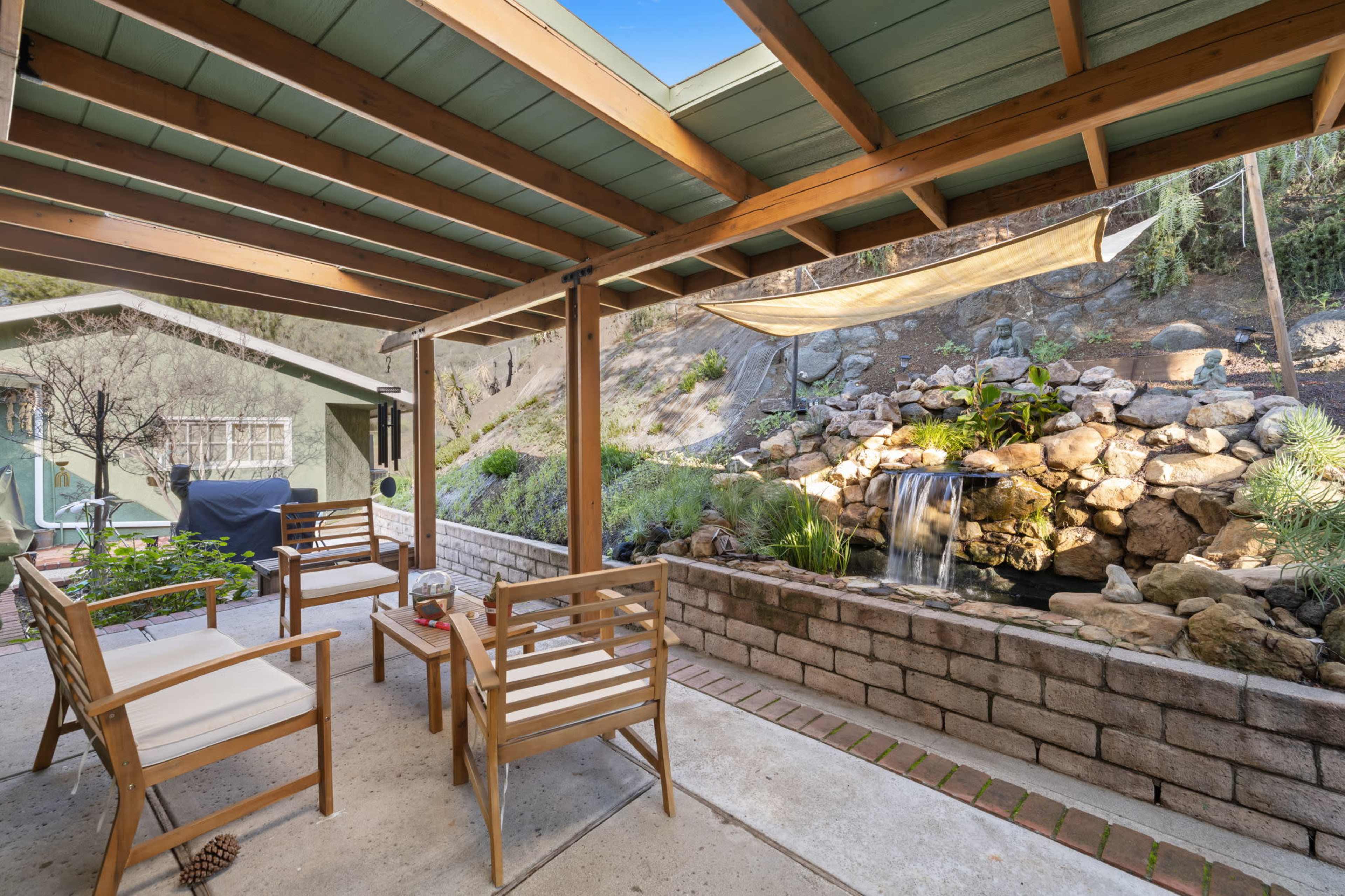 The image shows a covered patio area with wooden furniture, overlooking a rock garden with a small waterfall.