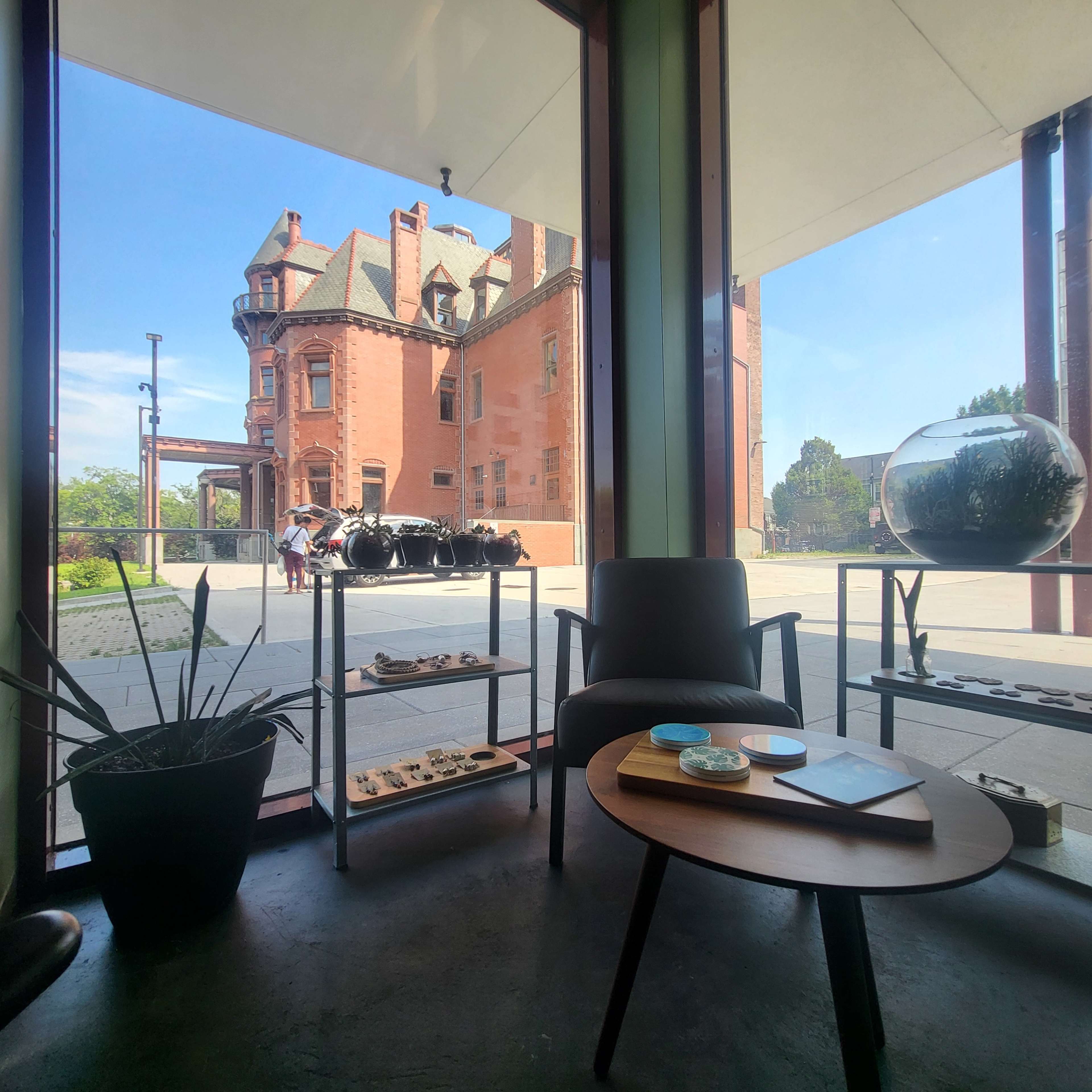 A modern interior space with a view of a historic brick building and outdoor seating, featuring a plant and display tables.