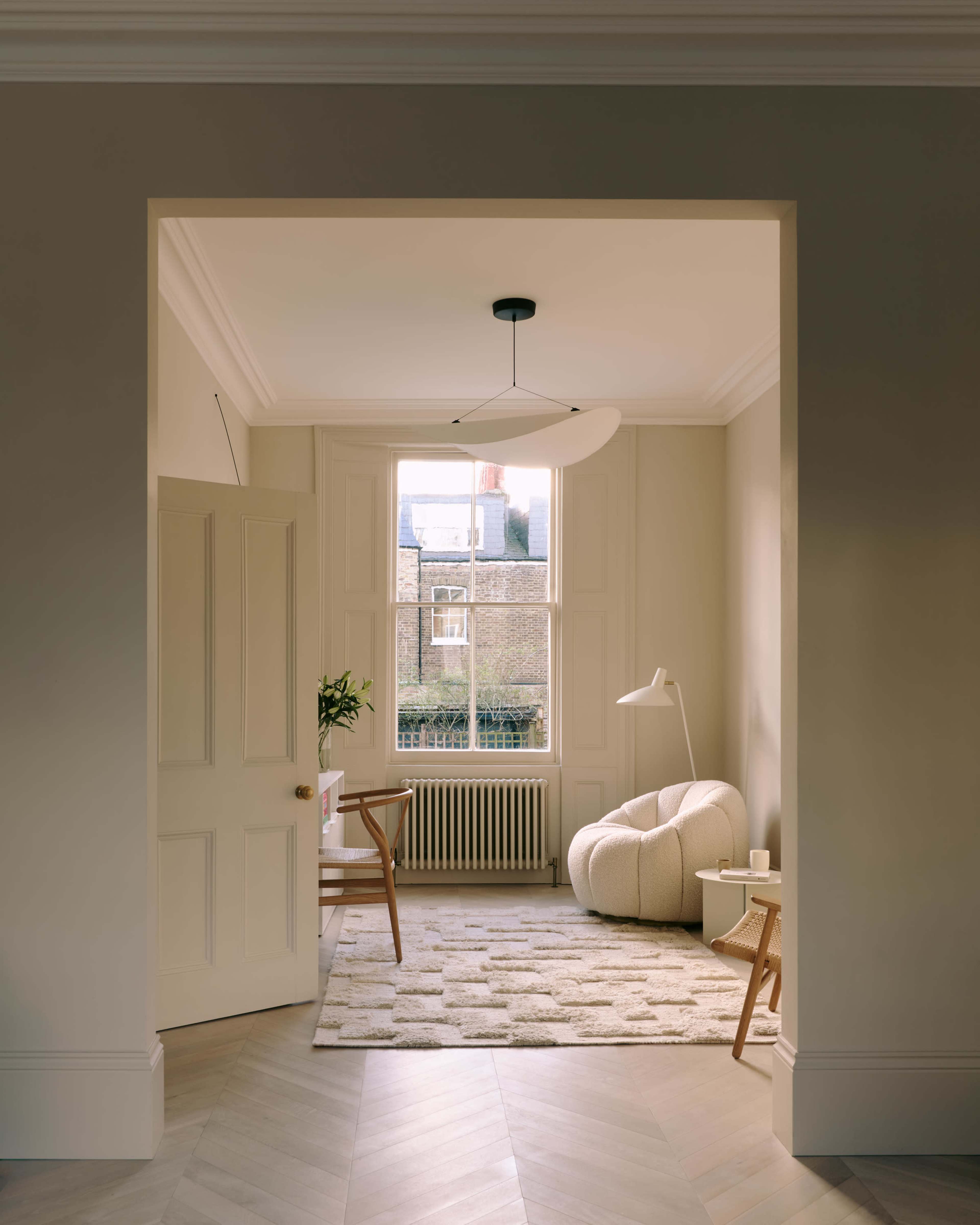 A beige room featuring a cozy seating area with a large round chair, a desk, and a floor lamp, illuminated by natural light from a window.