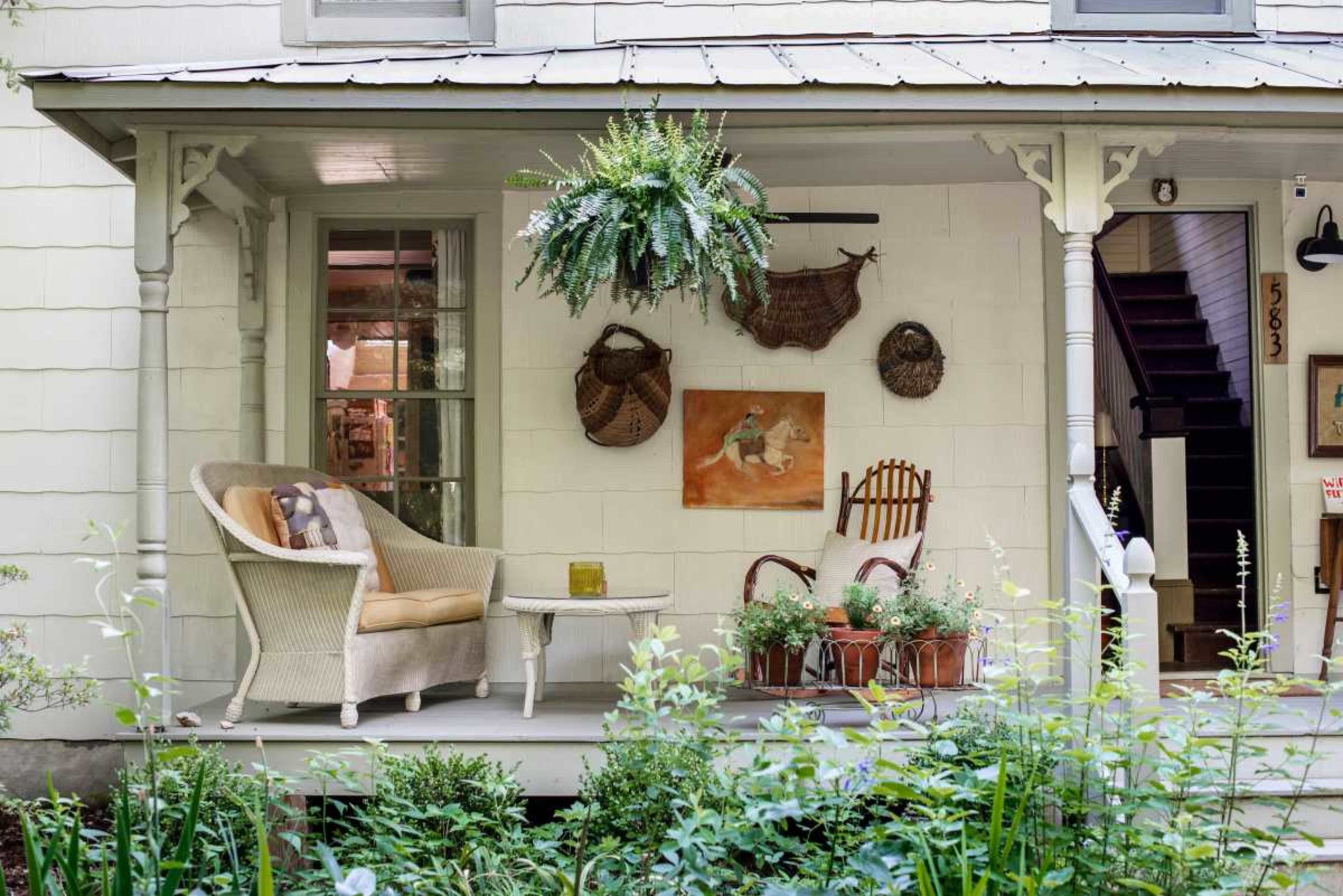 A front porch with a wicker chair, a small table, wall art, and potted plants surrounded by greenery.