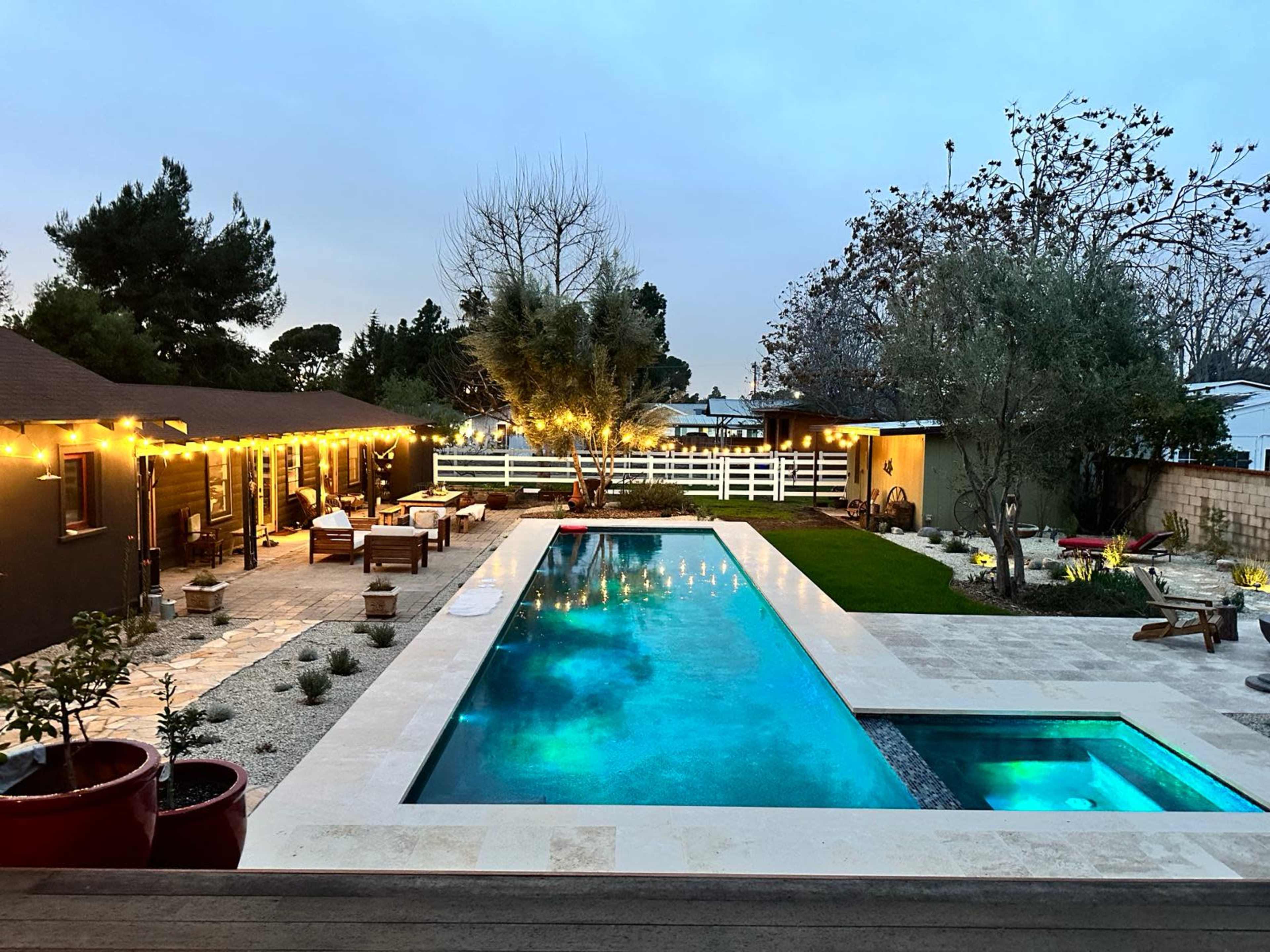 The image shows a backyard with a rectangular swimming pool surrounded by a patio, landscaped area, and string lights illuminating the space in the evening.