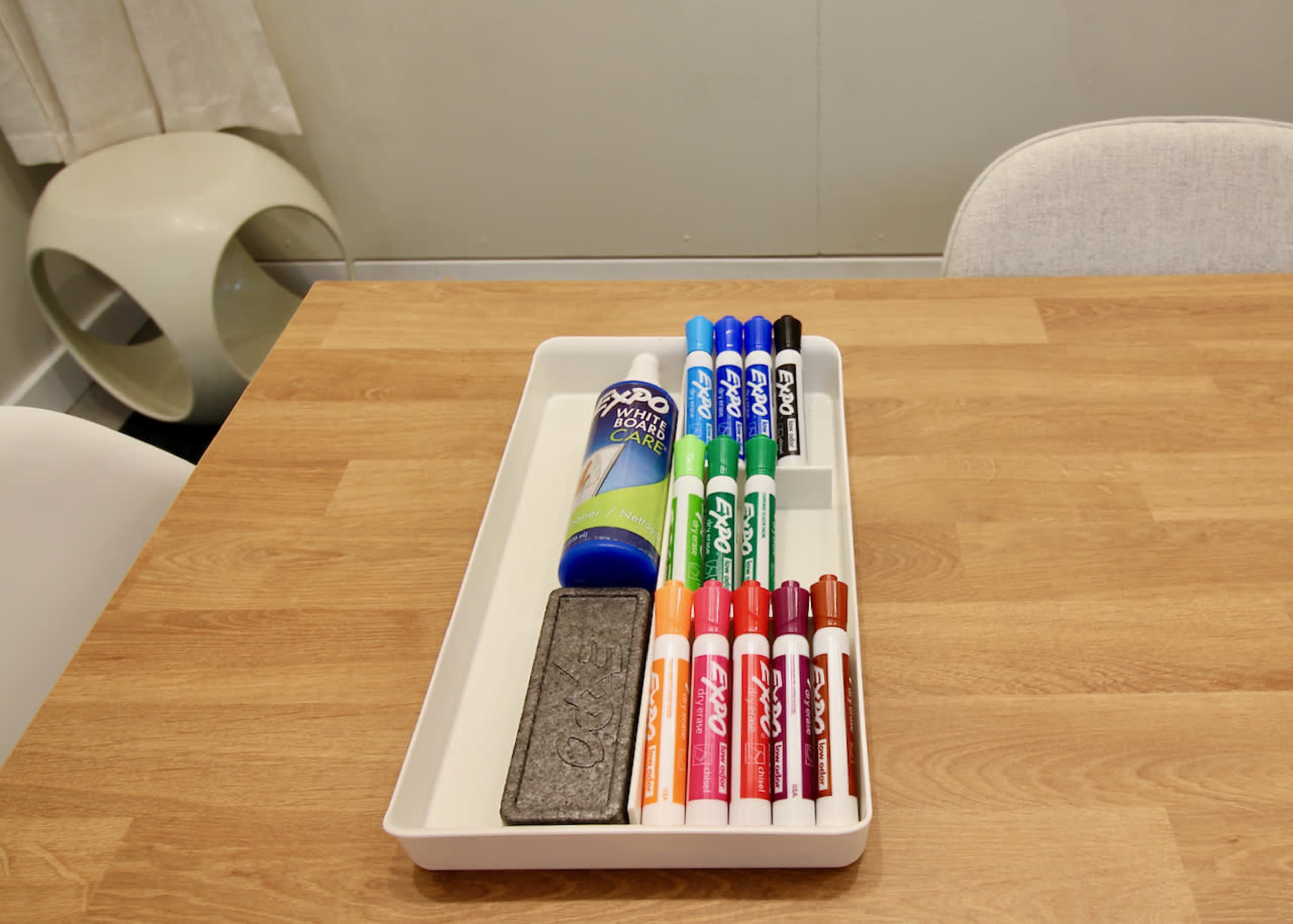 A tray on a wooden table holds various colored markers, a dry erase cleaner, and a gray eraser.