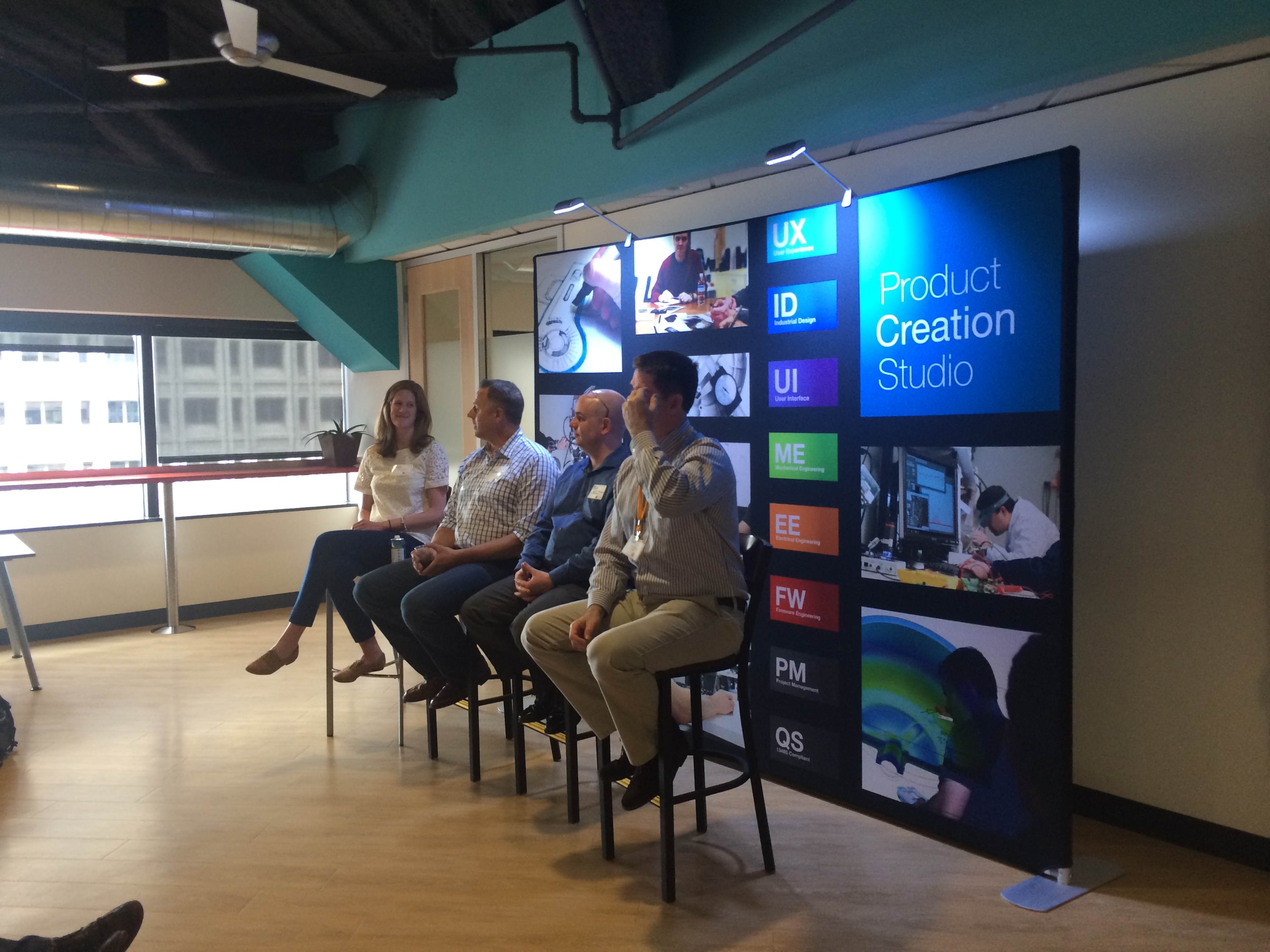 A panel of four speakers sits on stools in front of a display banner titled "Product Creation Studio" in a modern workspace.