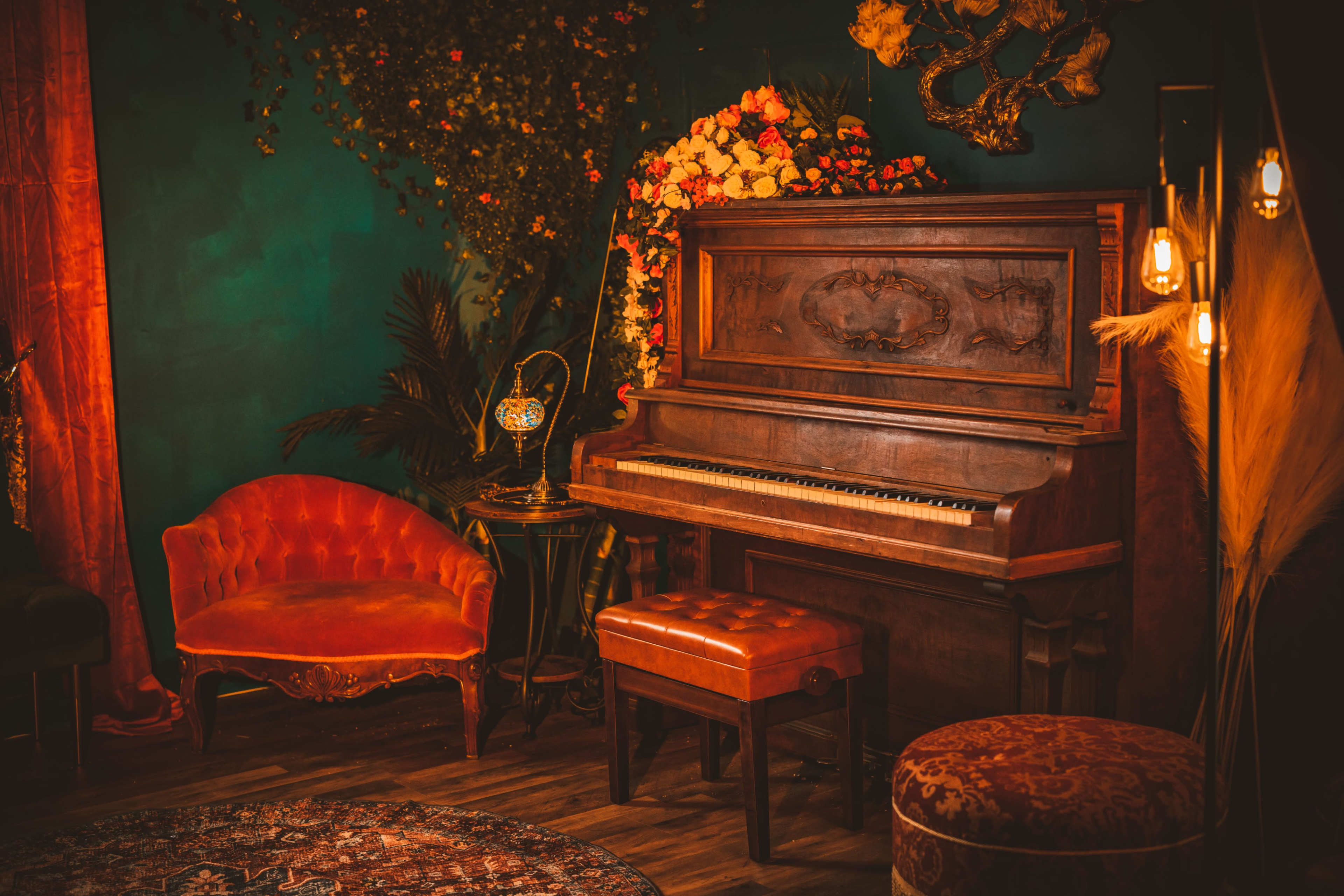 A vintage wooden piano sits next to an ornate orange couch in a warmly decorated room filled with flowers and soft lighting.