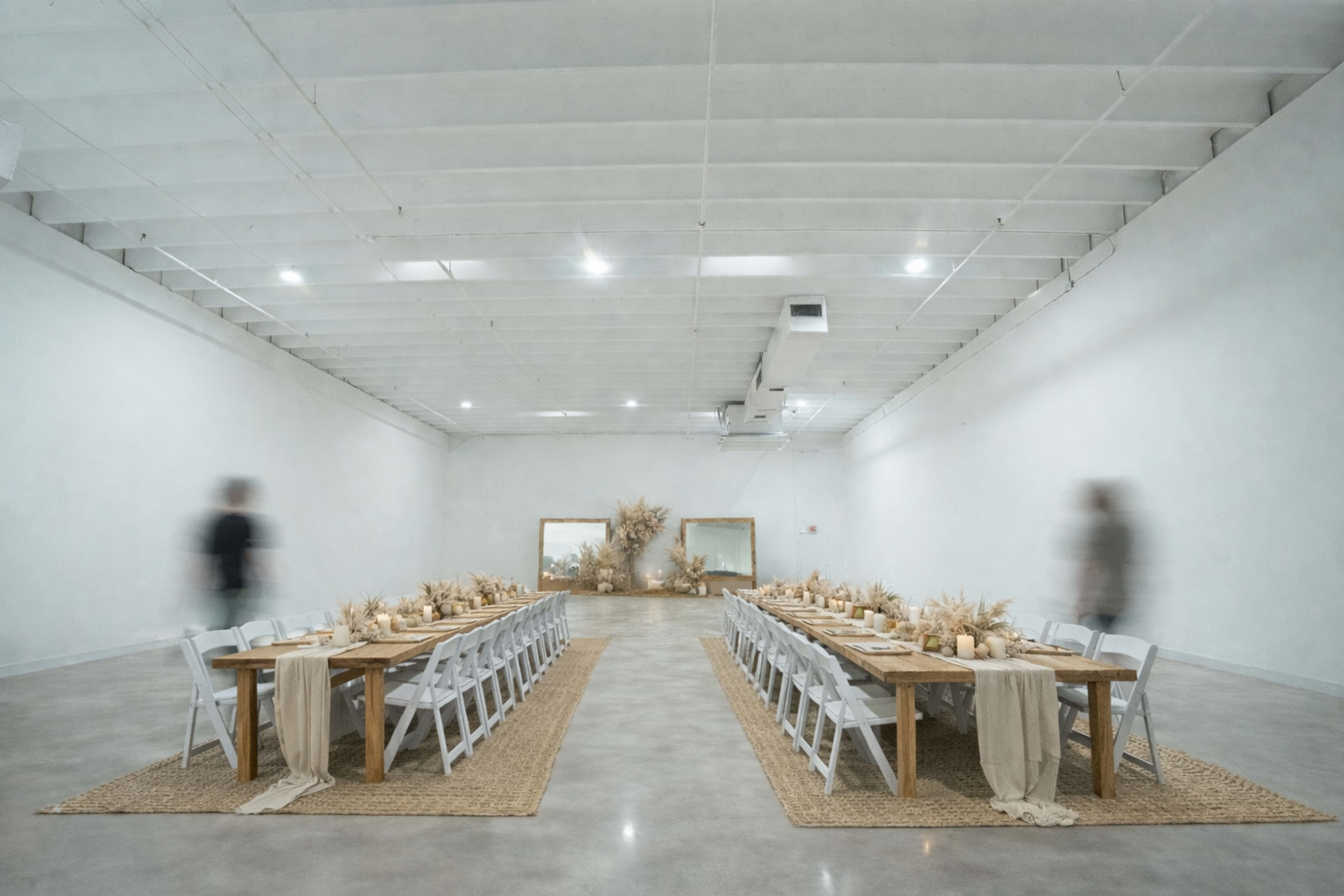 A spacious, empty event hall features two long banquet tables adorned with natural decor and is lined with white chairs, flanked by large mirrors on a plain wall.