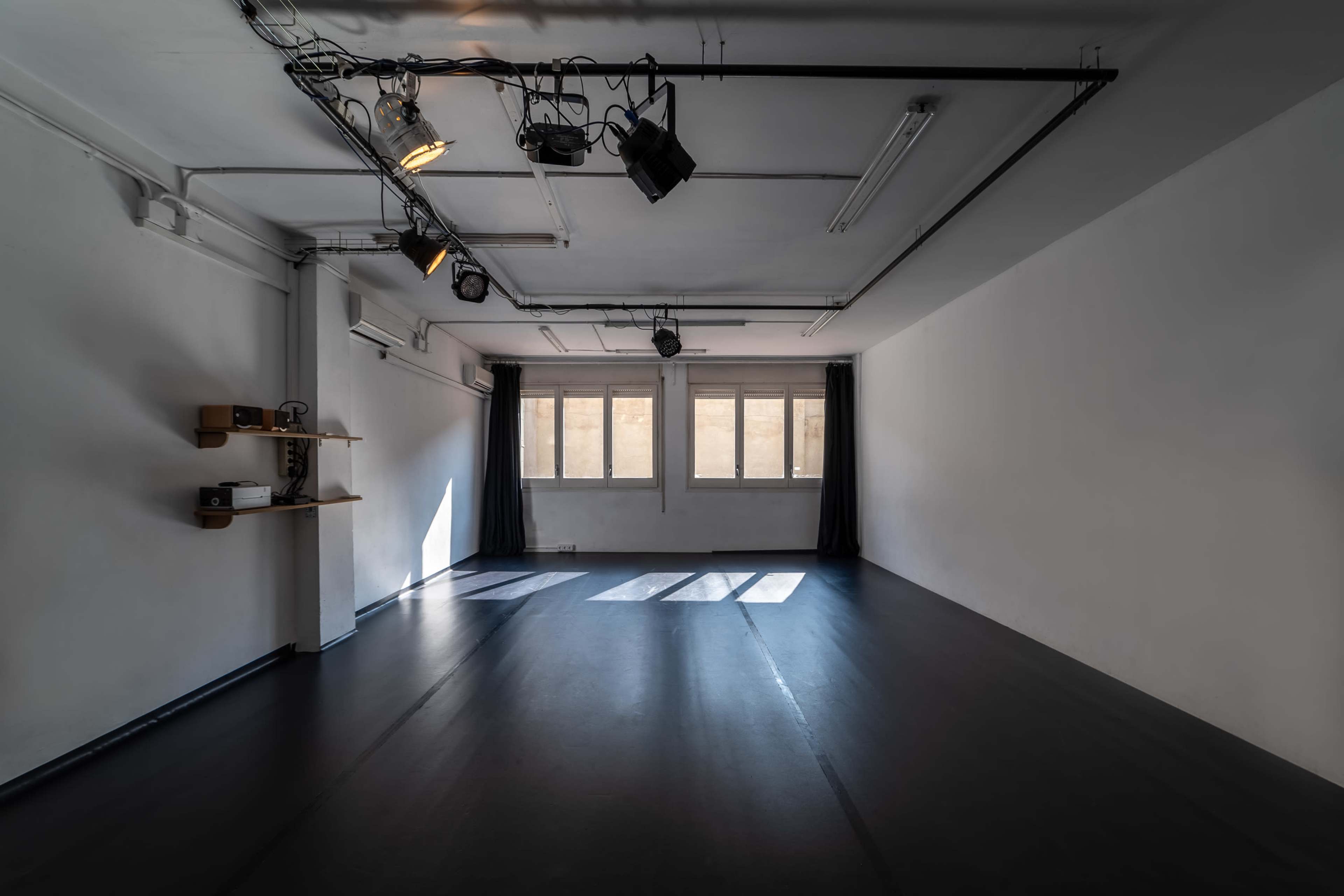An empty, well-lit studio space with gray walls, black flooring, and daylight streaming through large windows.
