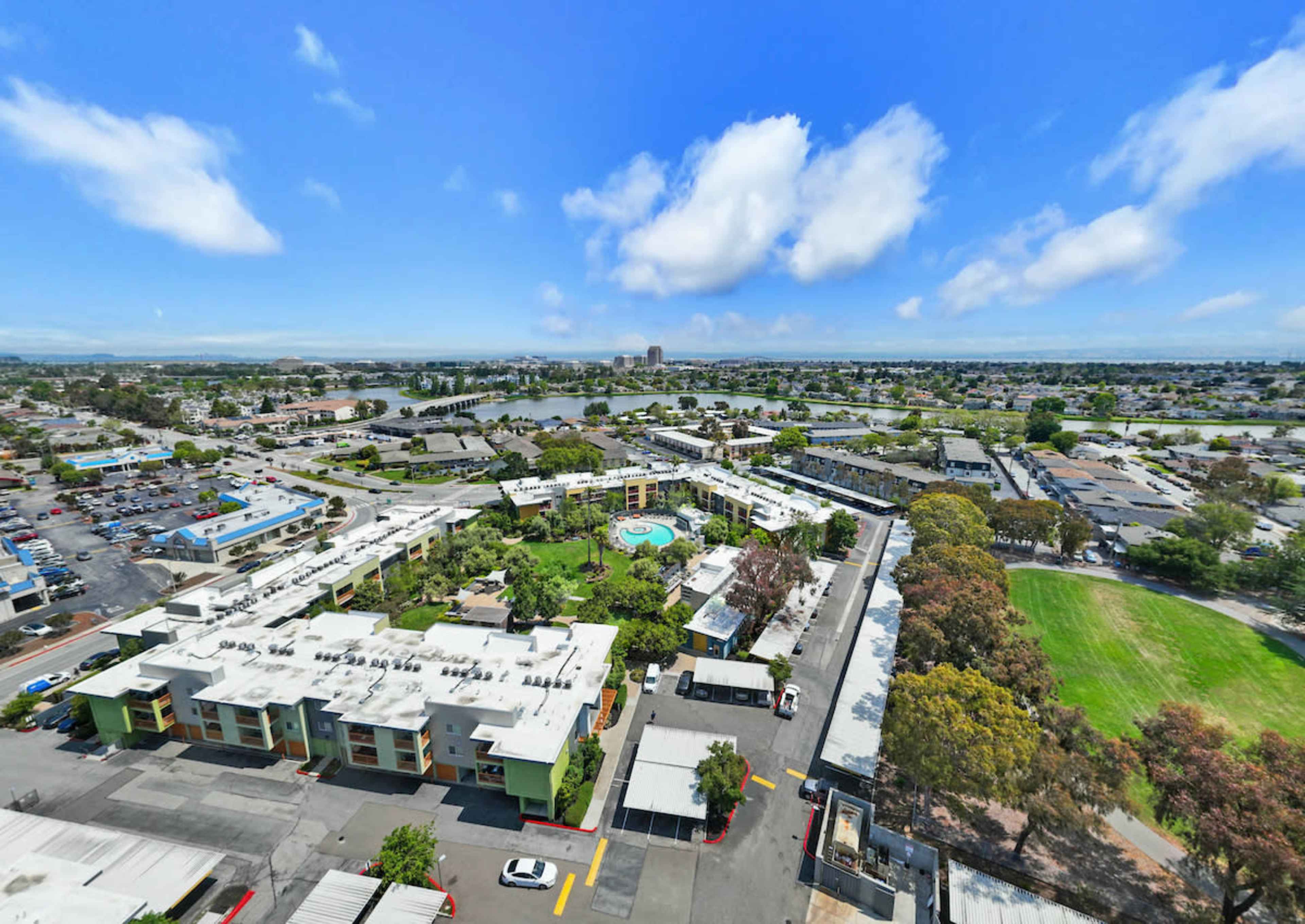 A wide aerial view of a suburban area featuring a swimming pool surrounded by residential buildings, parking lots, and green spaces.