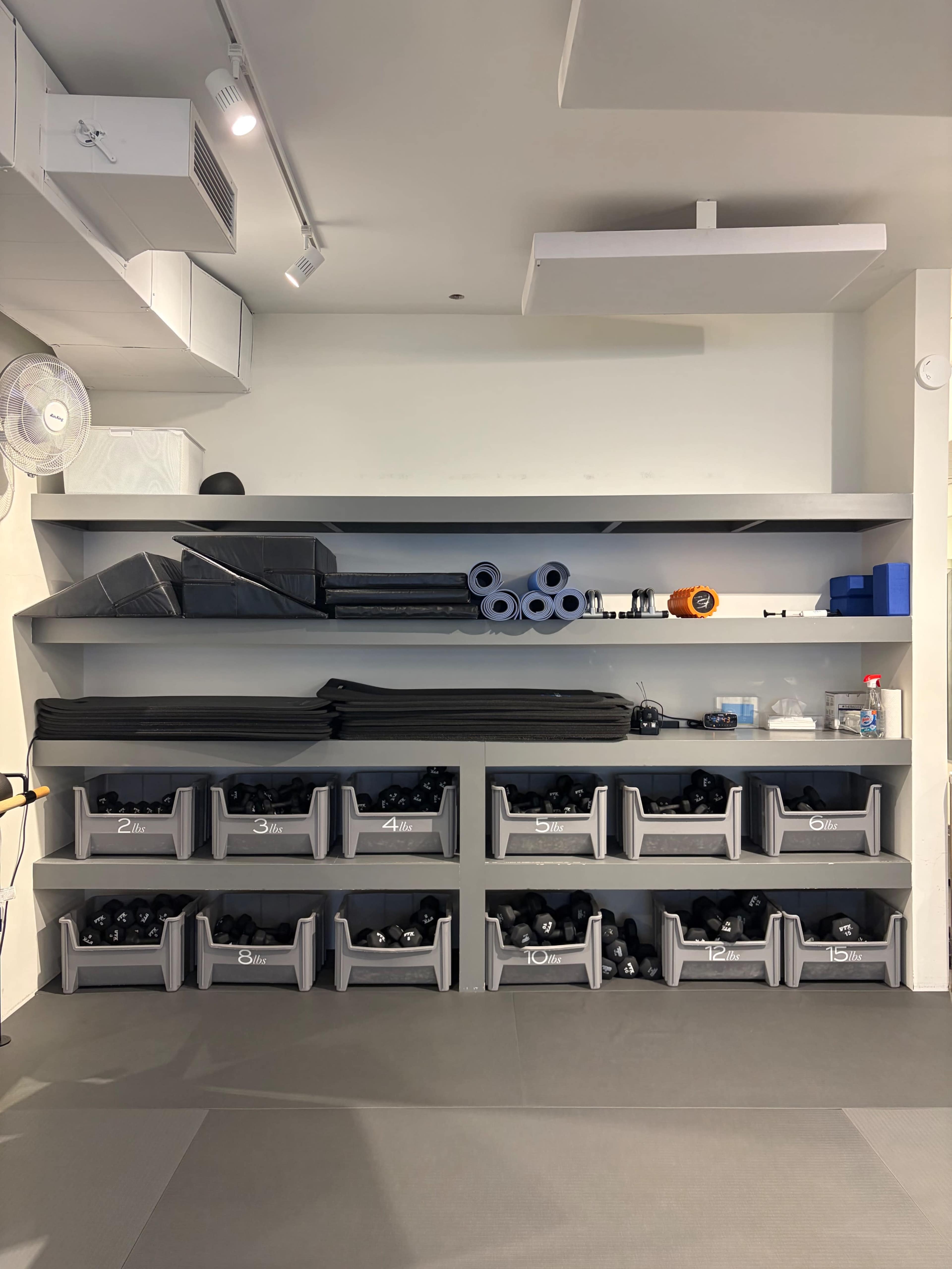The image shows a minimalist fitness space with shelves holding various exercise equipment, including foam rollers, mats, and organized bins filled with dumbbells.
