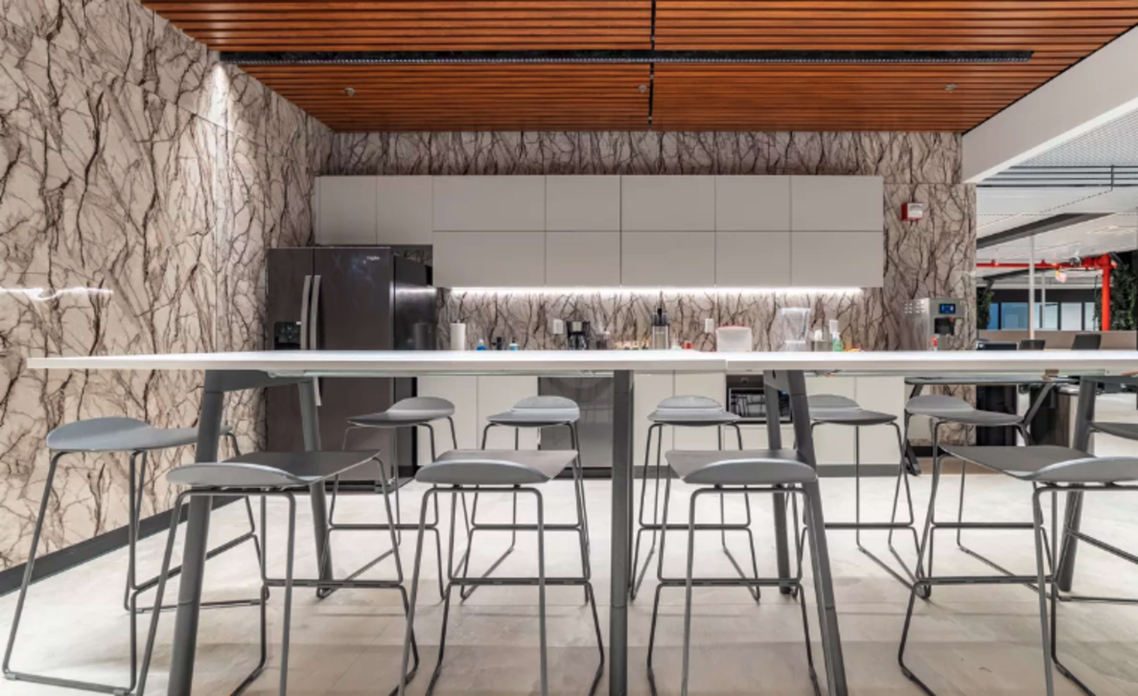 A modern kitchen with a large white countertop, gray stools, and a marble-patterned wall, featuring appliances and minimalist cabinetry.