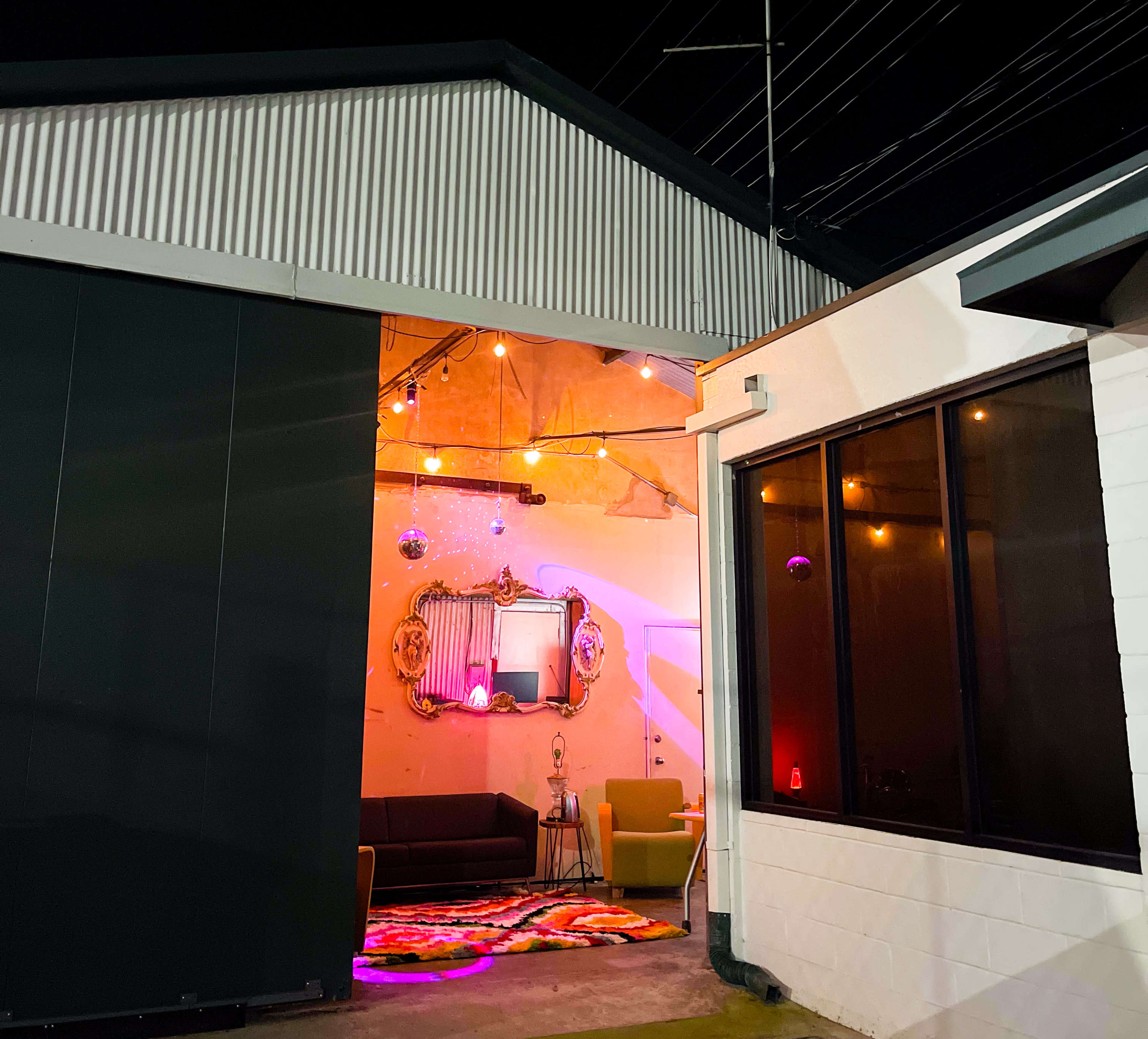 A modern lounge area viewed through an open entrance, illuminated by colorful lights and featuring a couch, chairs, and decorative elements.