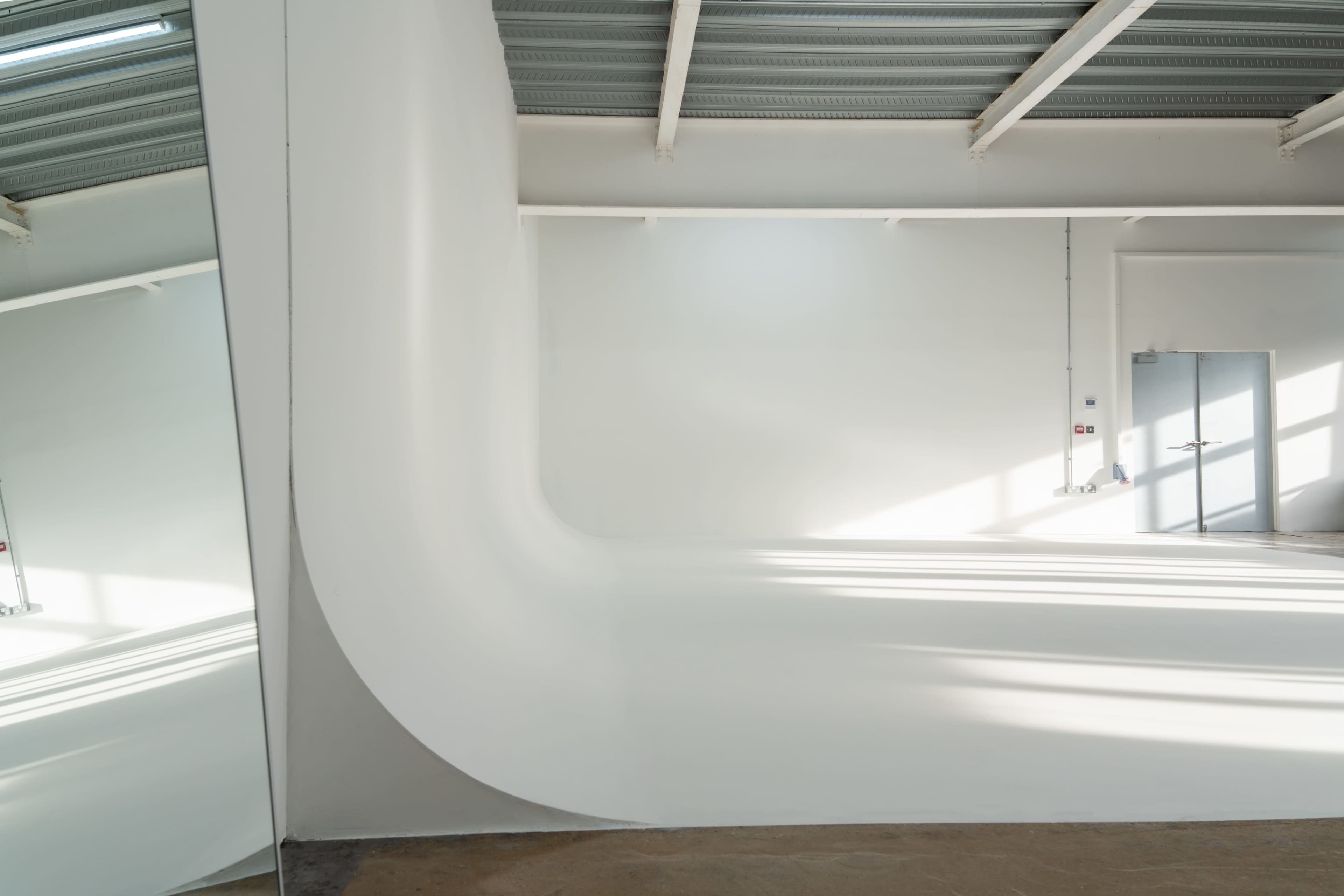 The image shows a minimalist interior space with a curved white wall and ample natural light coming through a large window.