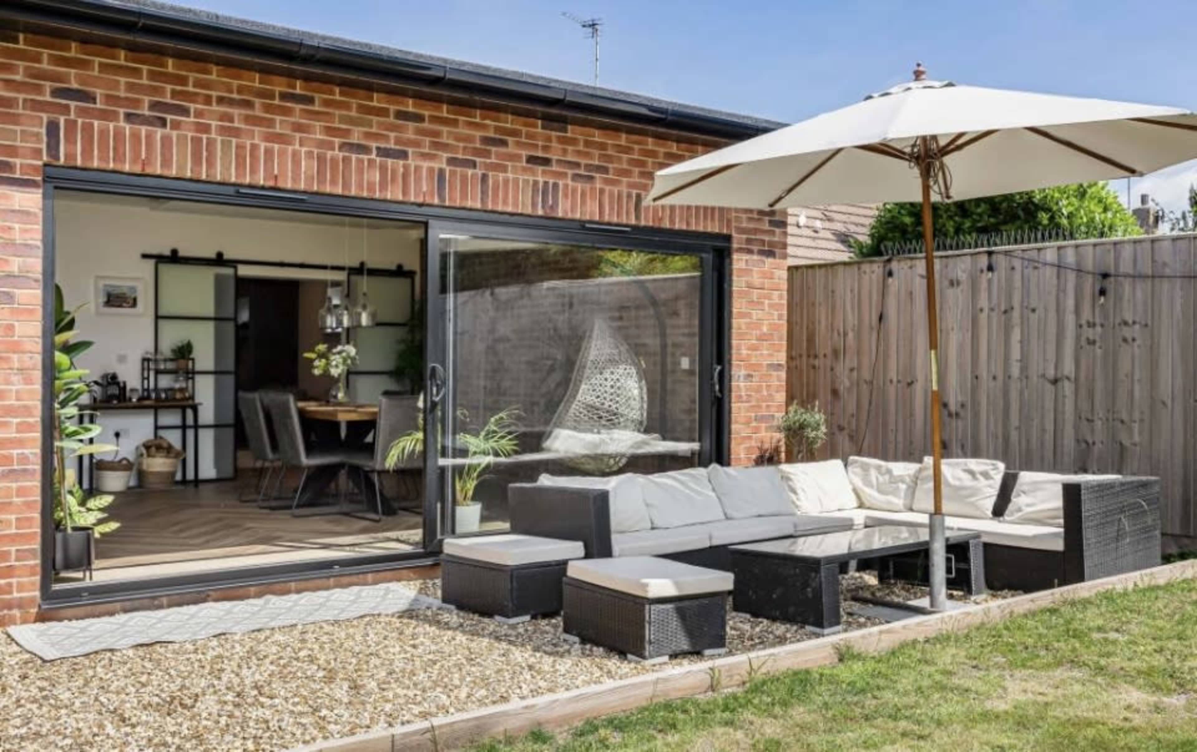 A modern patio area features a sectional sofa, a glass table, and a large umbrella, bordered by a gravel lawn and surrounded by brick walls.