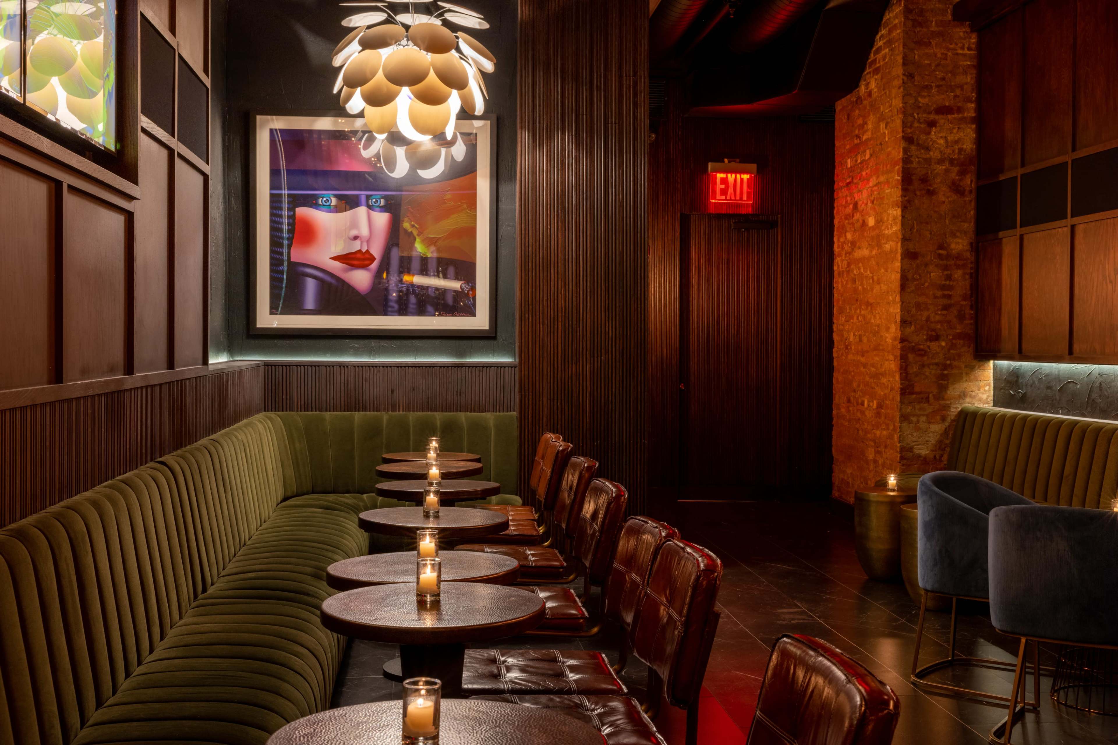 The image shows a dimly lit lounge area with brown leather chairs, green upholstered seating, small round tables, and a large artwork on the wall.