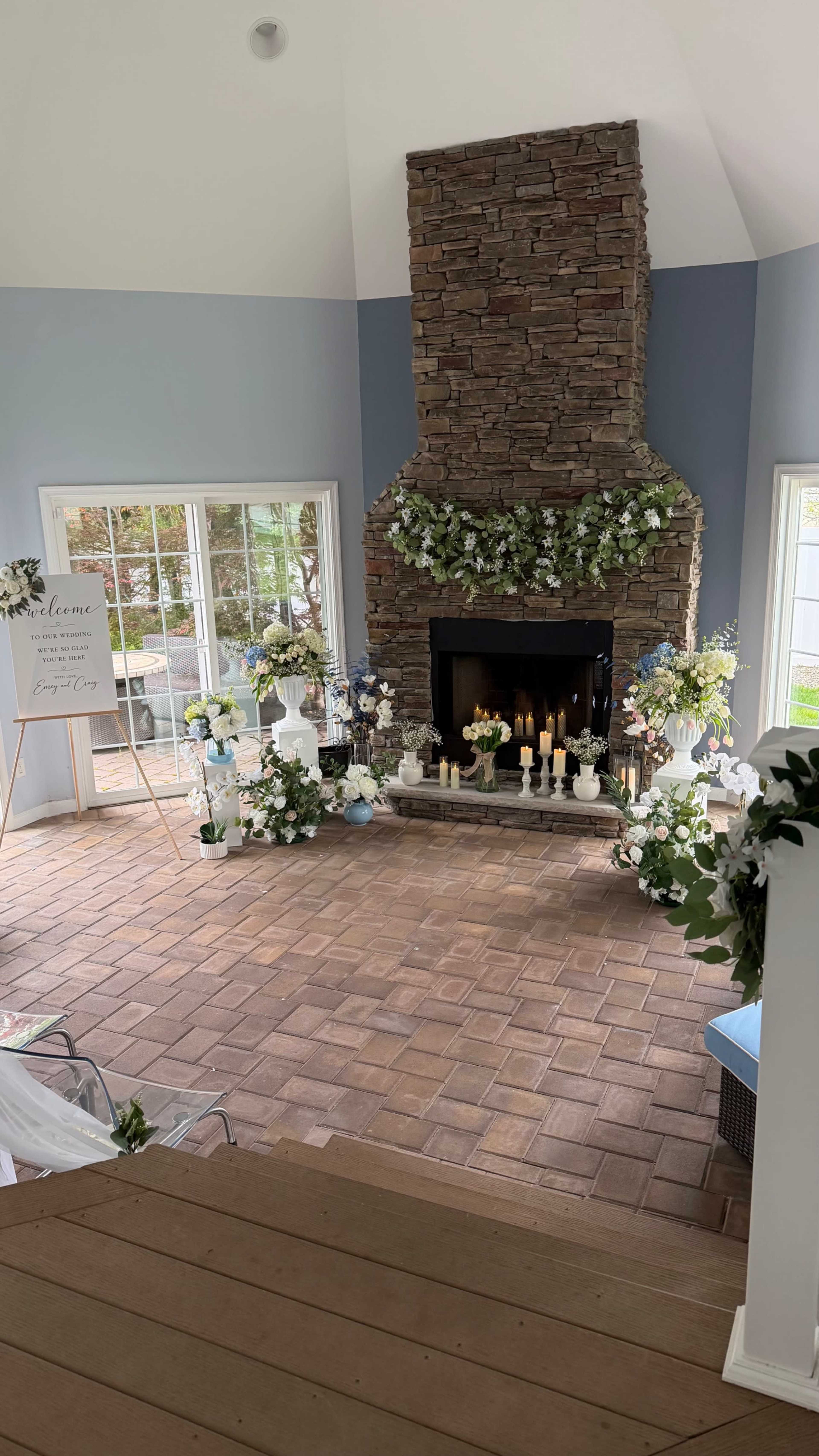 The image shows a spacious room with a stone fireplace adorned with greenery and floral arrangements, featuring large windows that offer a view of the outdoors.