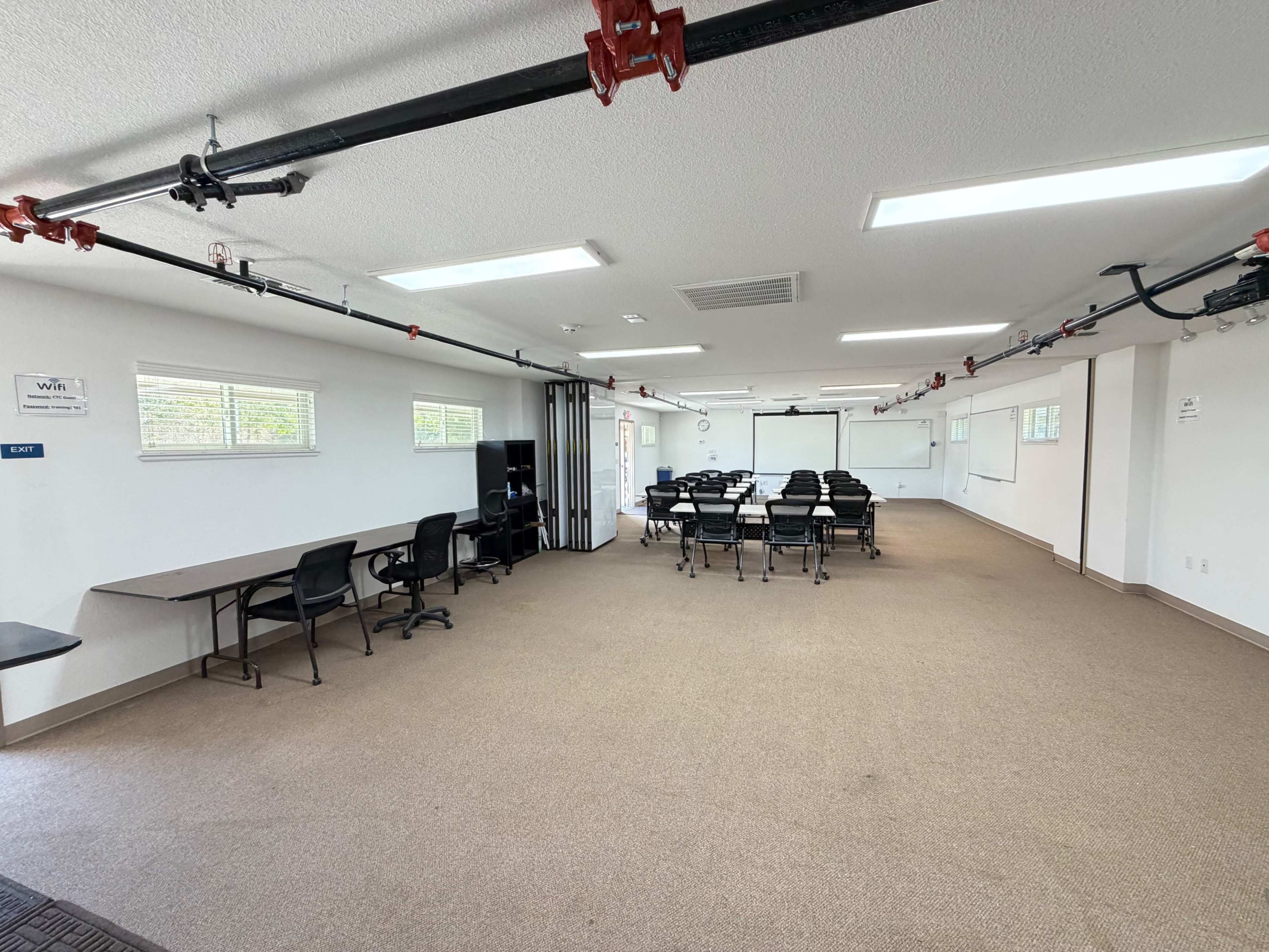 The image shows a spacious, well-lit meeting room featuring a long table with chairs arranged around it, along with a few desks and multiple windows.