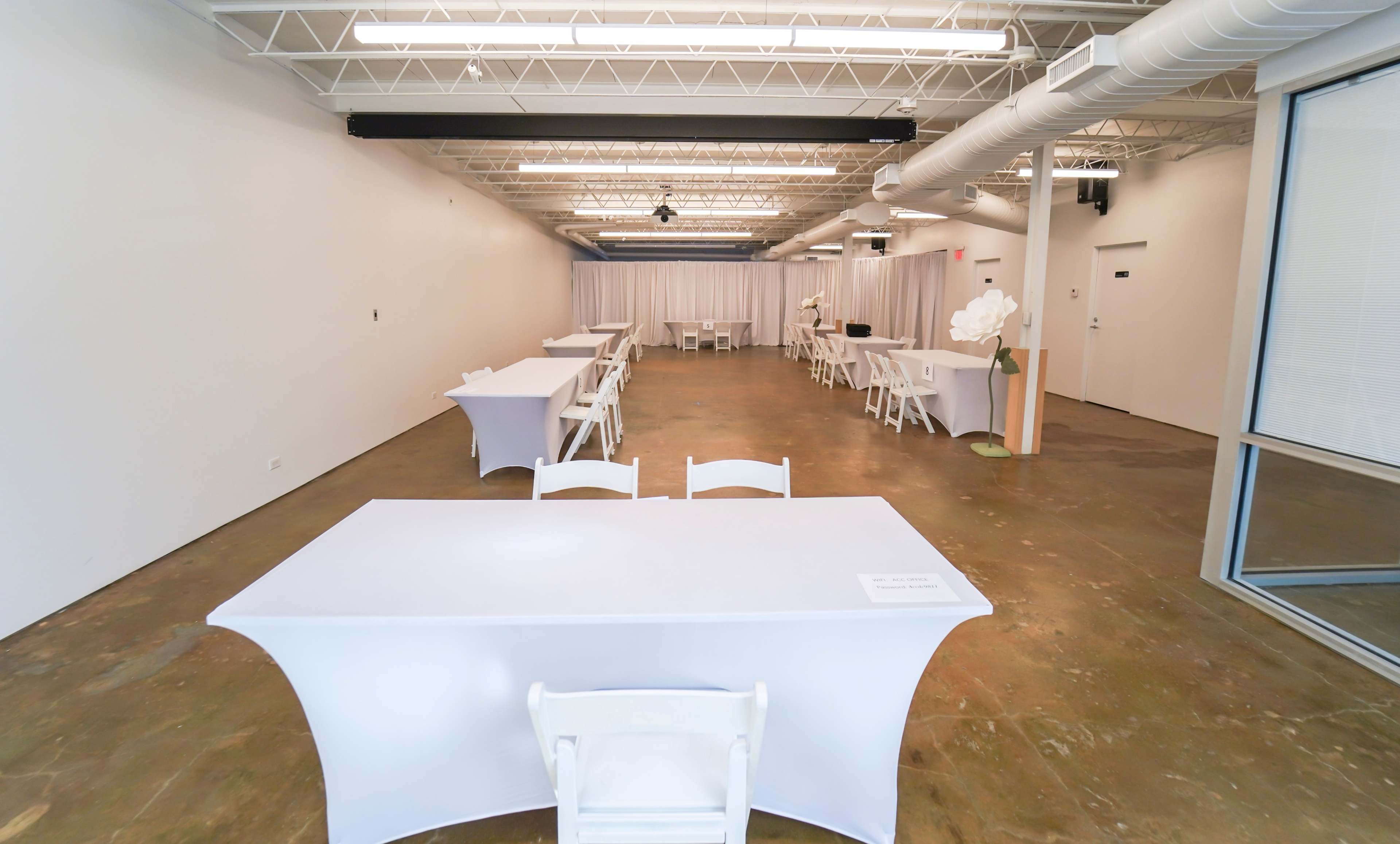 A spacious, empty room with white tables and chairs arranged neatly throughout, featuring a concrete floor and white walls.