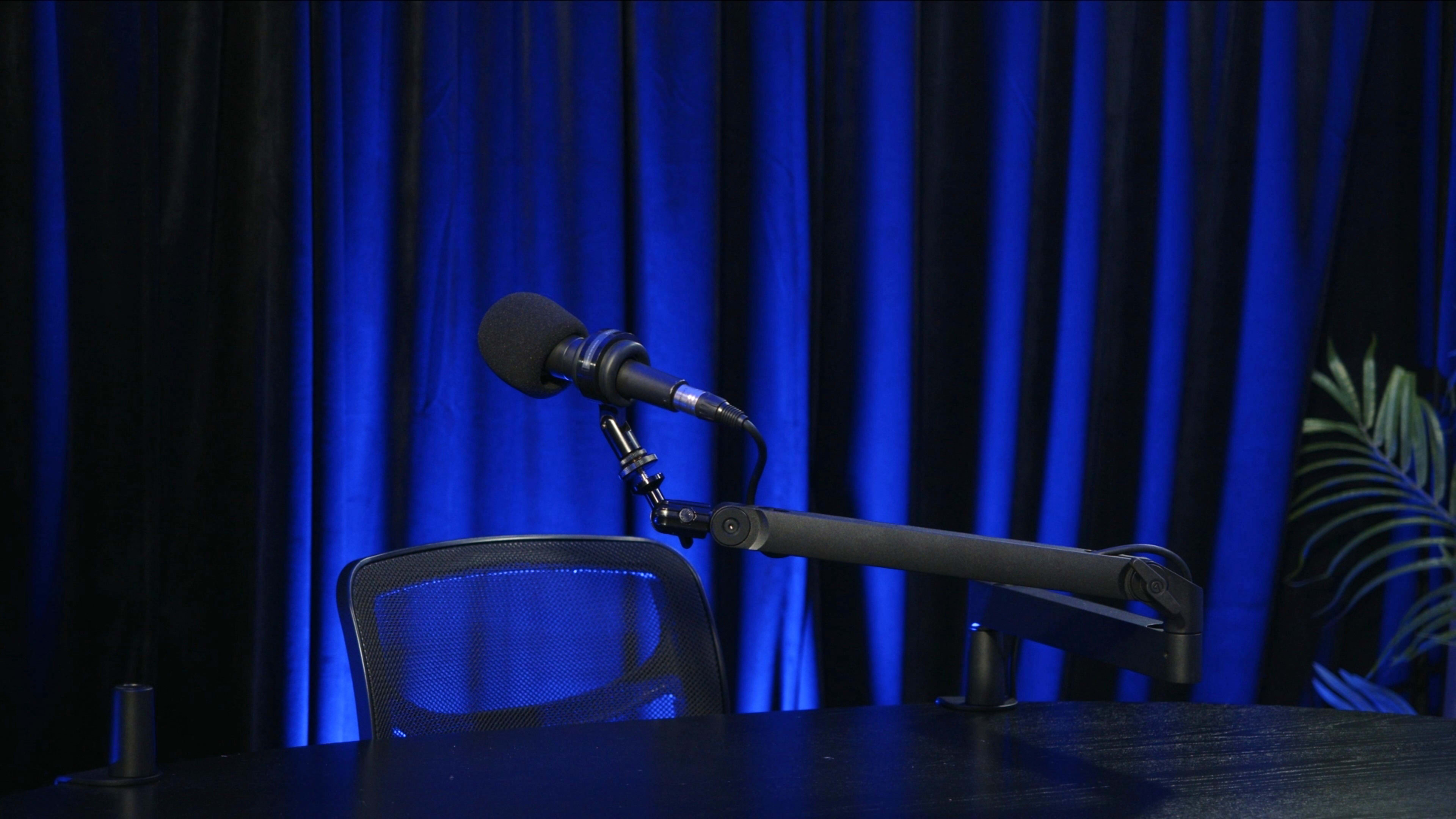 A microphone is mounted on a stand next to an empty chair in front of a table, with a backdrop of blue draping.