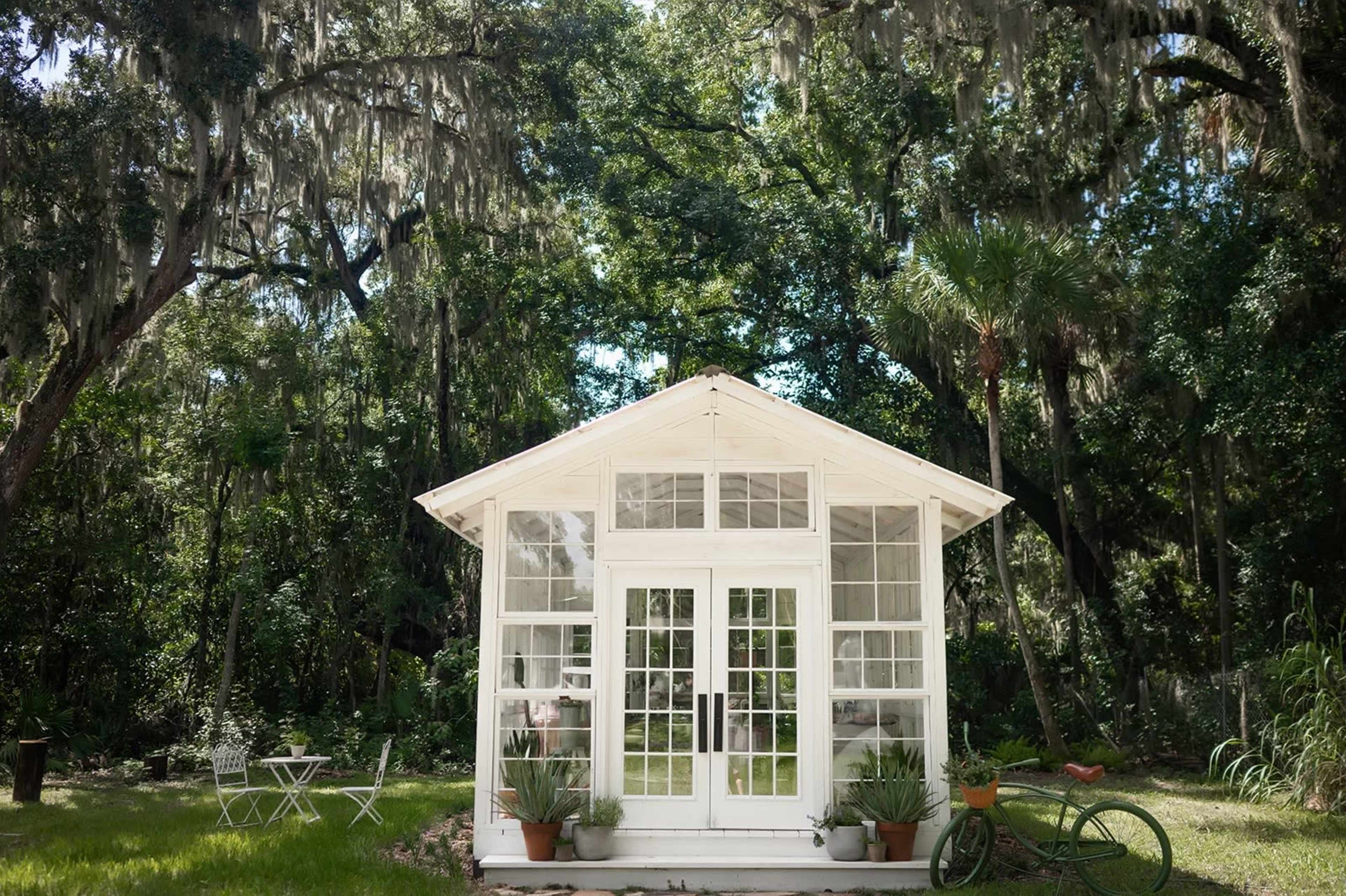 A white garden shed with large windows sits in a grassy area surrounded by trees.